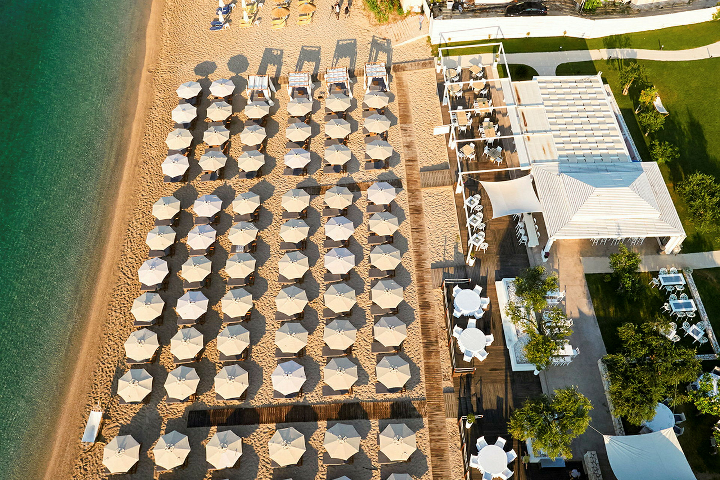 Flygfoto av en strand med uppradade solstolar och parasoller samt en restaurang med sittplatser. (AI-genererad text)