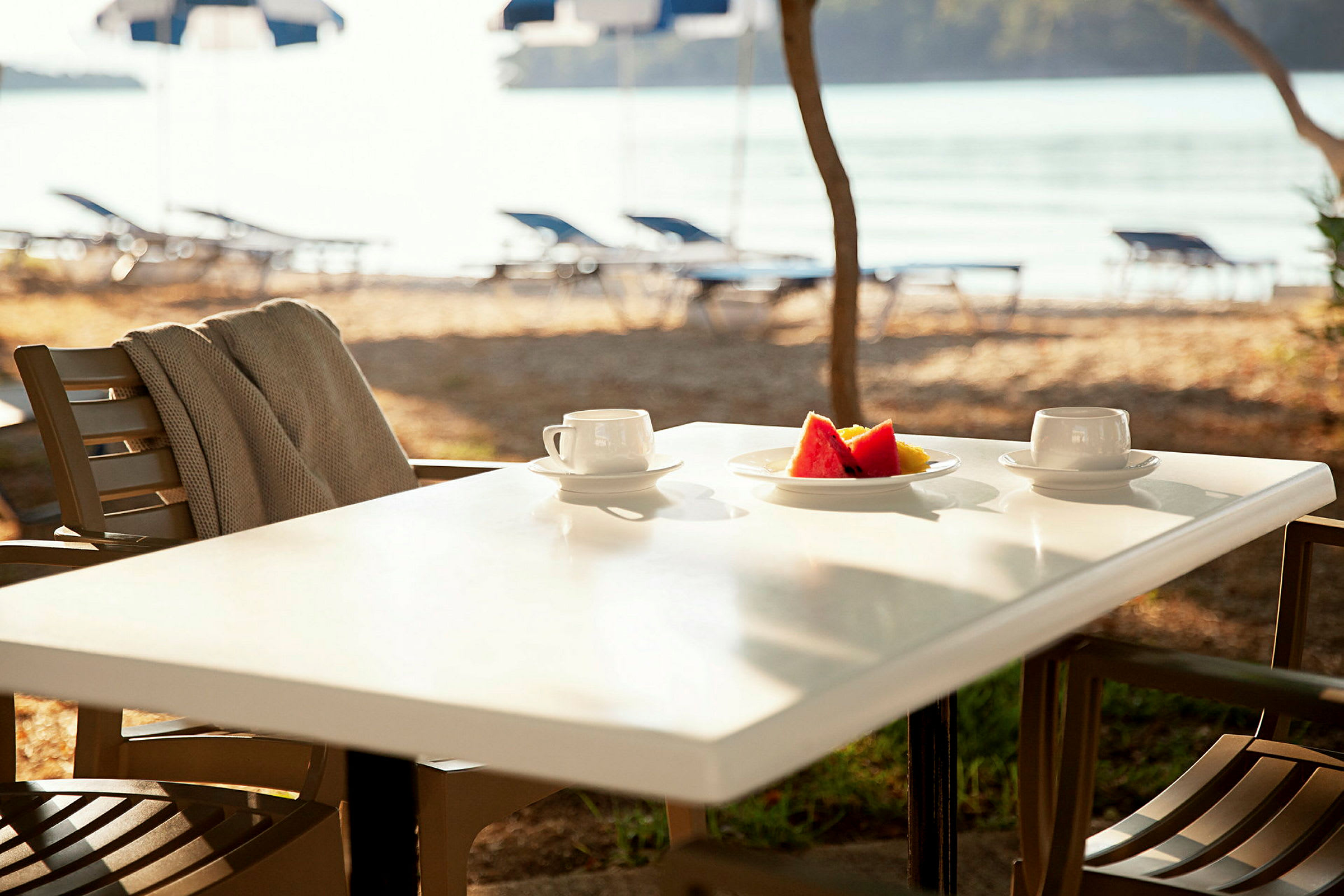 En utomhusservering med ett bord med två kaffekoppar och en tallrik med frukt. Bakgrunden visar strand och vatten. (AI-genererad text)