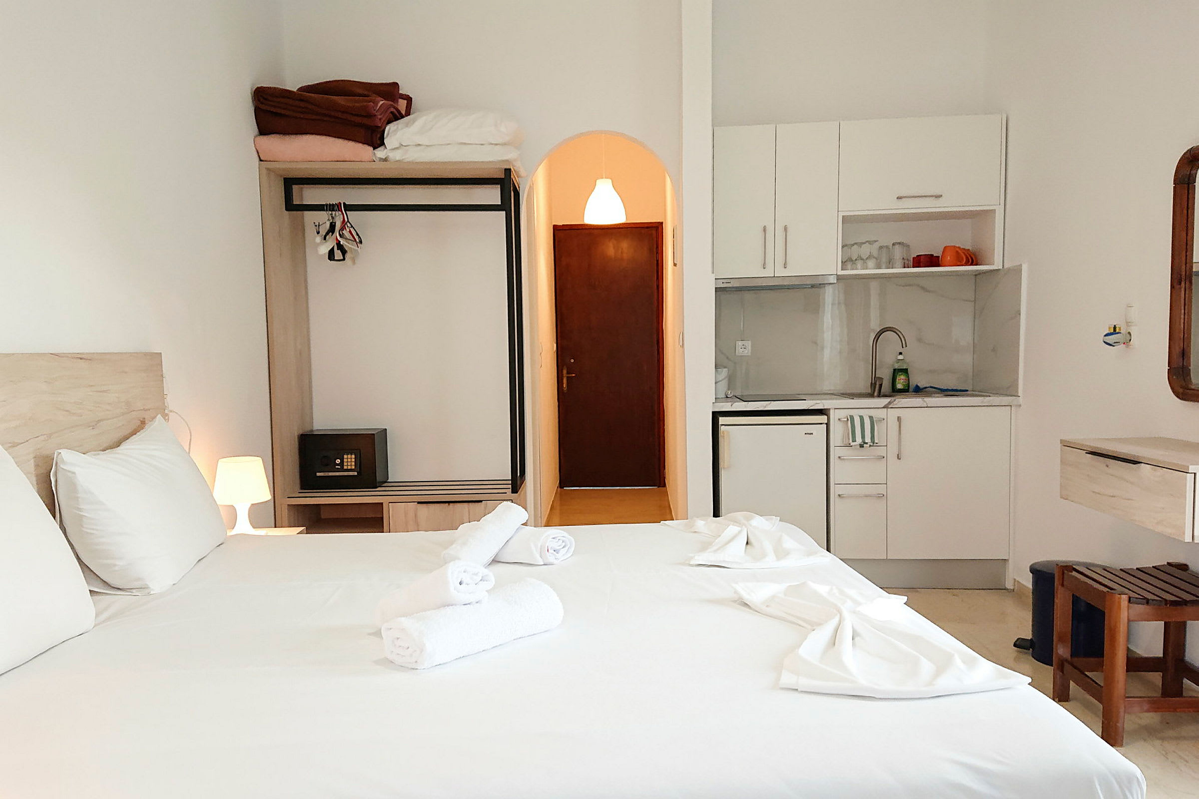 Hotellrum med en bäddad dubbelsäng, garderob, dörr, kokvrå och stol. (AI-genererad text)