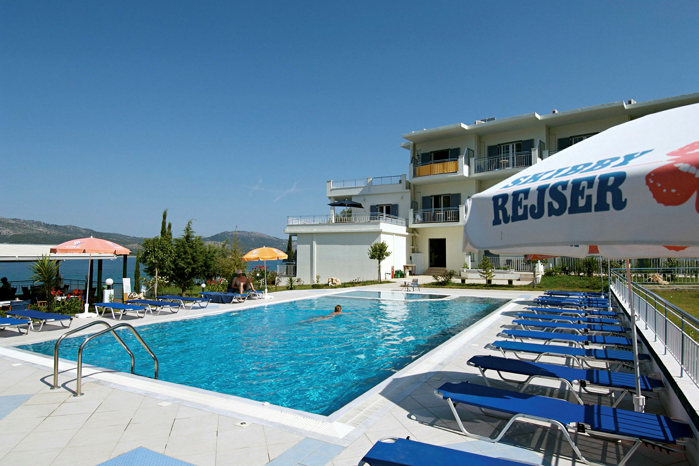 Bild av en pool med blå solstolar och parasoller framför ett hotell. (AI-genererad text)