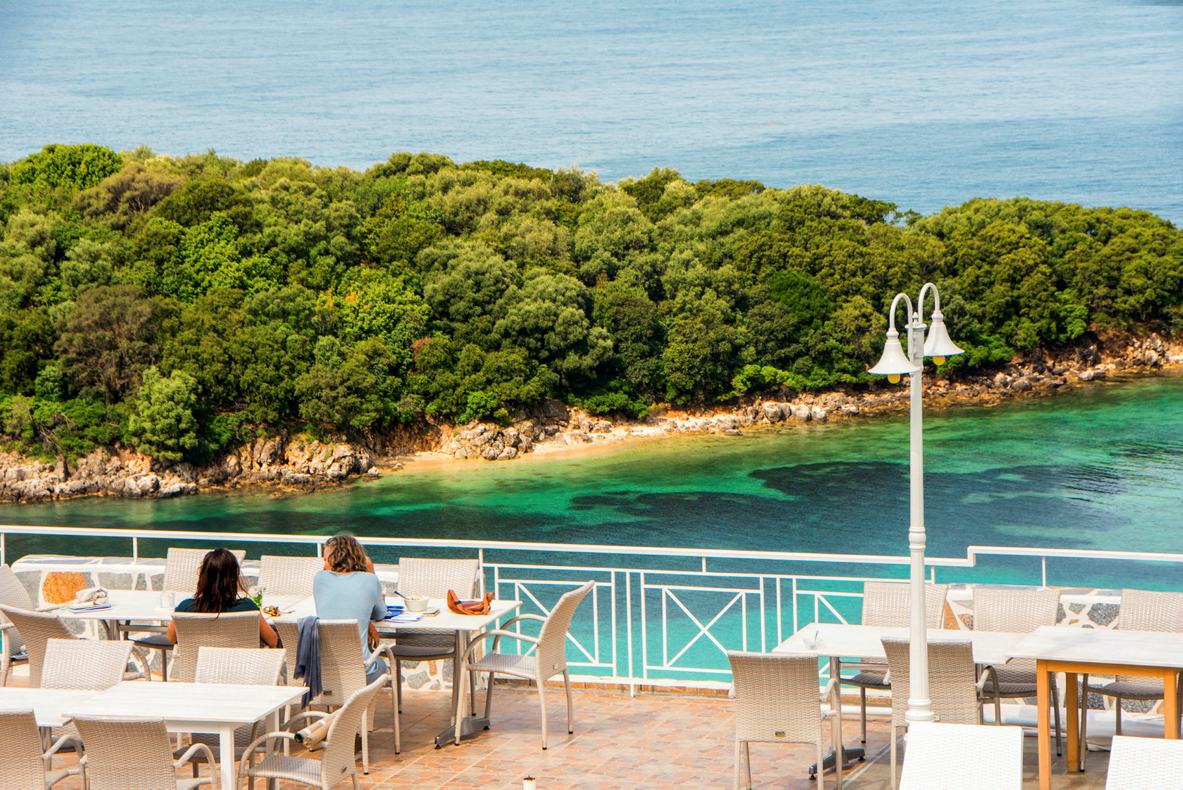 Två personer sitter vid bord på en terrass med utsikt över havet och en grönskande kustlinje. (AI-genererad text)