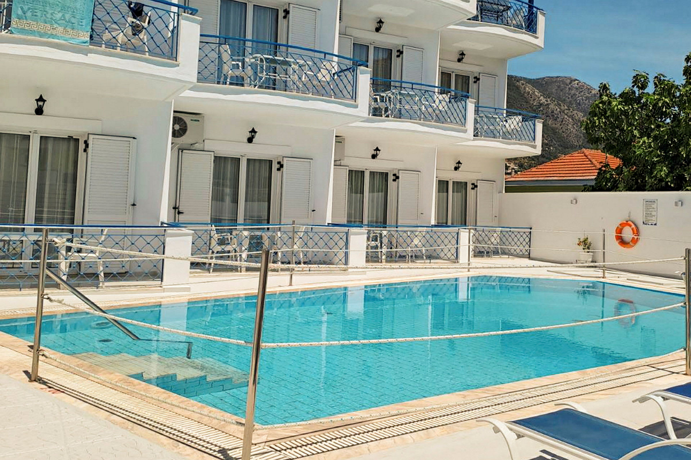 Bild av en pool framför ett vitmålat hotell med blåa balkonger och solstolar vid poolen. (AI-genererad text)