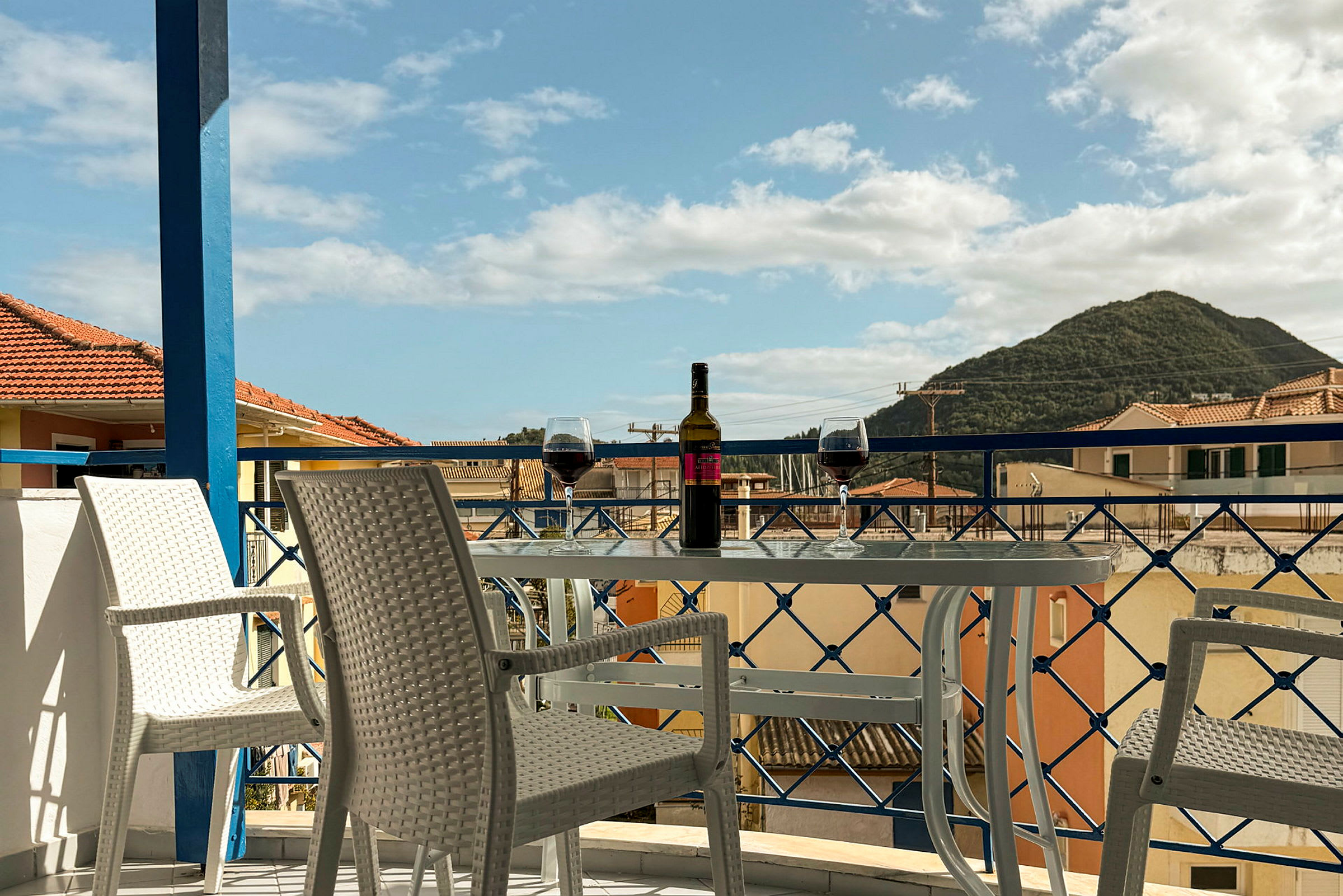 Utsikt från en balkong med bord, två glas vin och en vinflaska, samt stolar. I bakgrunden syns hustak och en grönskande kulle. (AI-genererad text)
