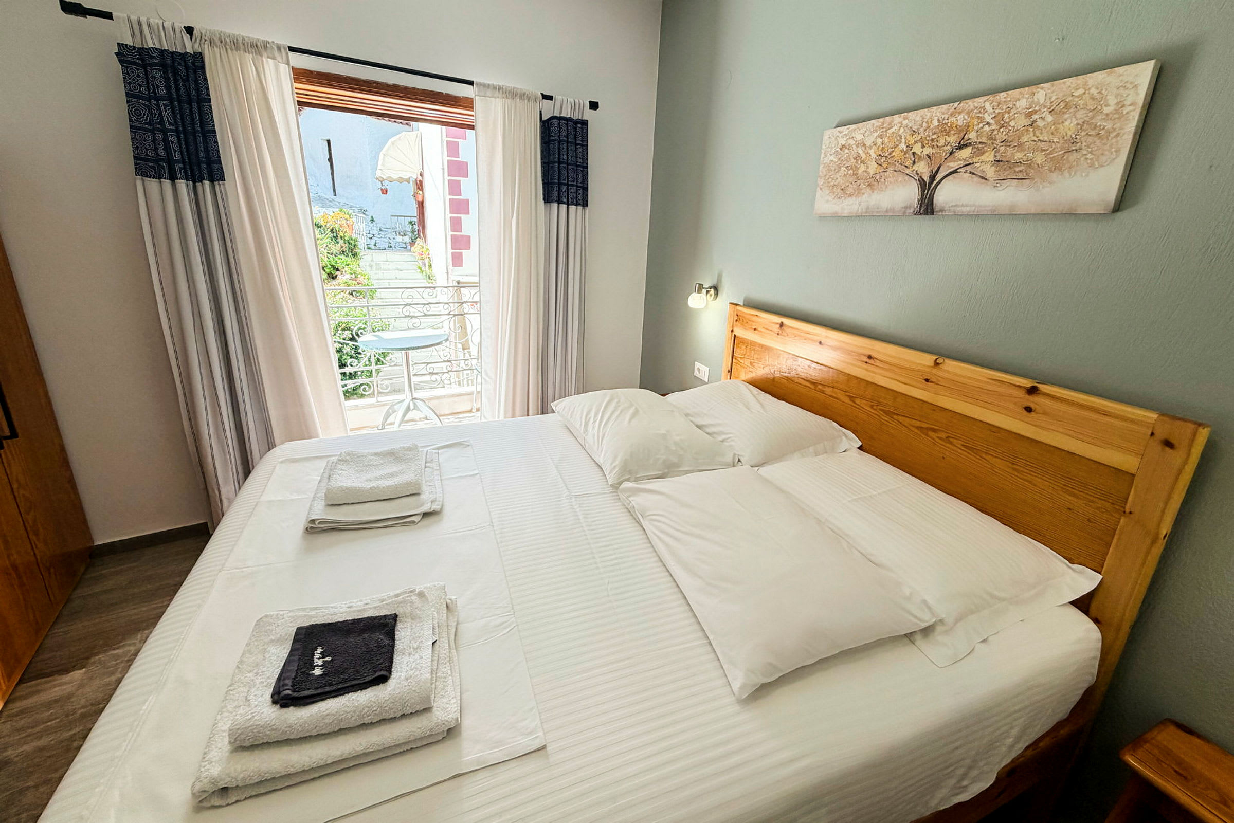 Hotellrum med dubbelsäng, vit sänglinne och trädesign på sänggavel. Balkong med utsikt kan ses genom glasade dörrar. (AI-genererad text)