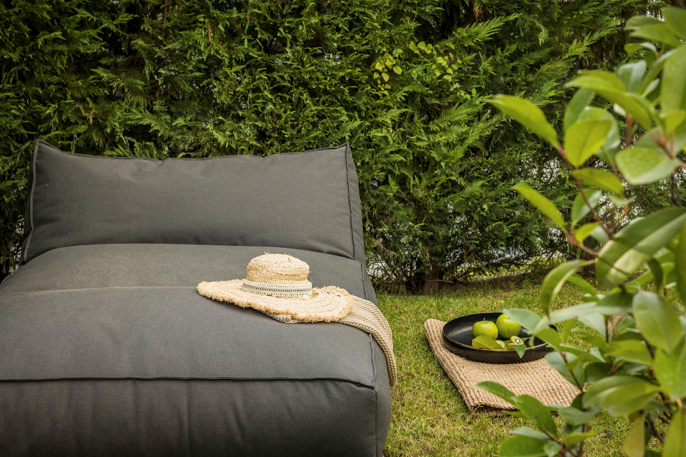 Grå sittpuff med halmhatt och filt, vid sidan ligger gröna äpplen i en skål på en matta, omgiven av grönska. (AI-genererad text)