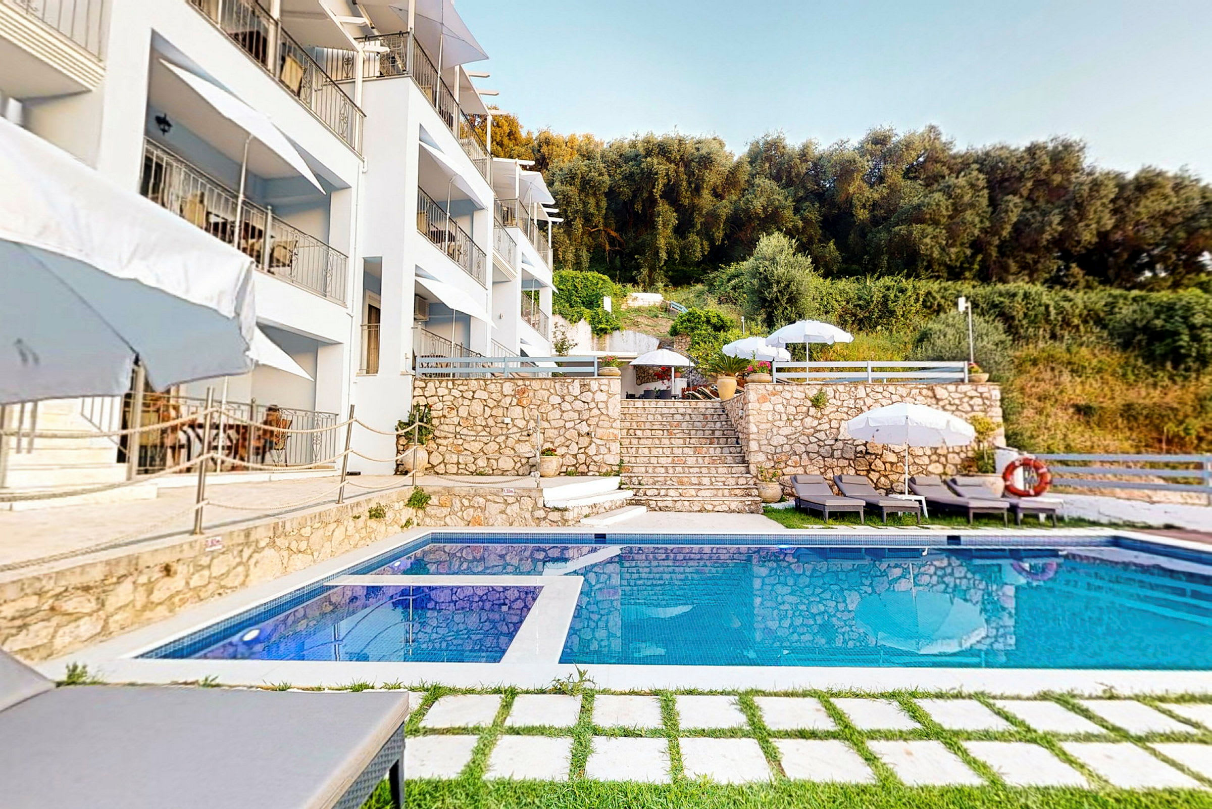 En pool framför en byggnad, omgivet av parasoller, solsängar och grönska. (AI-genererad text)