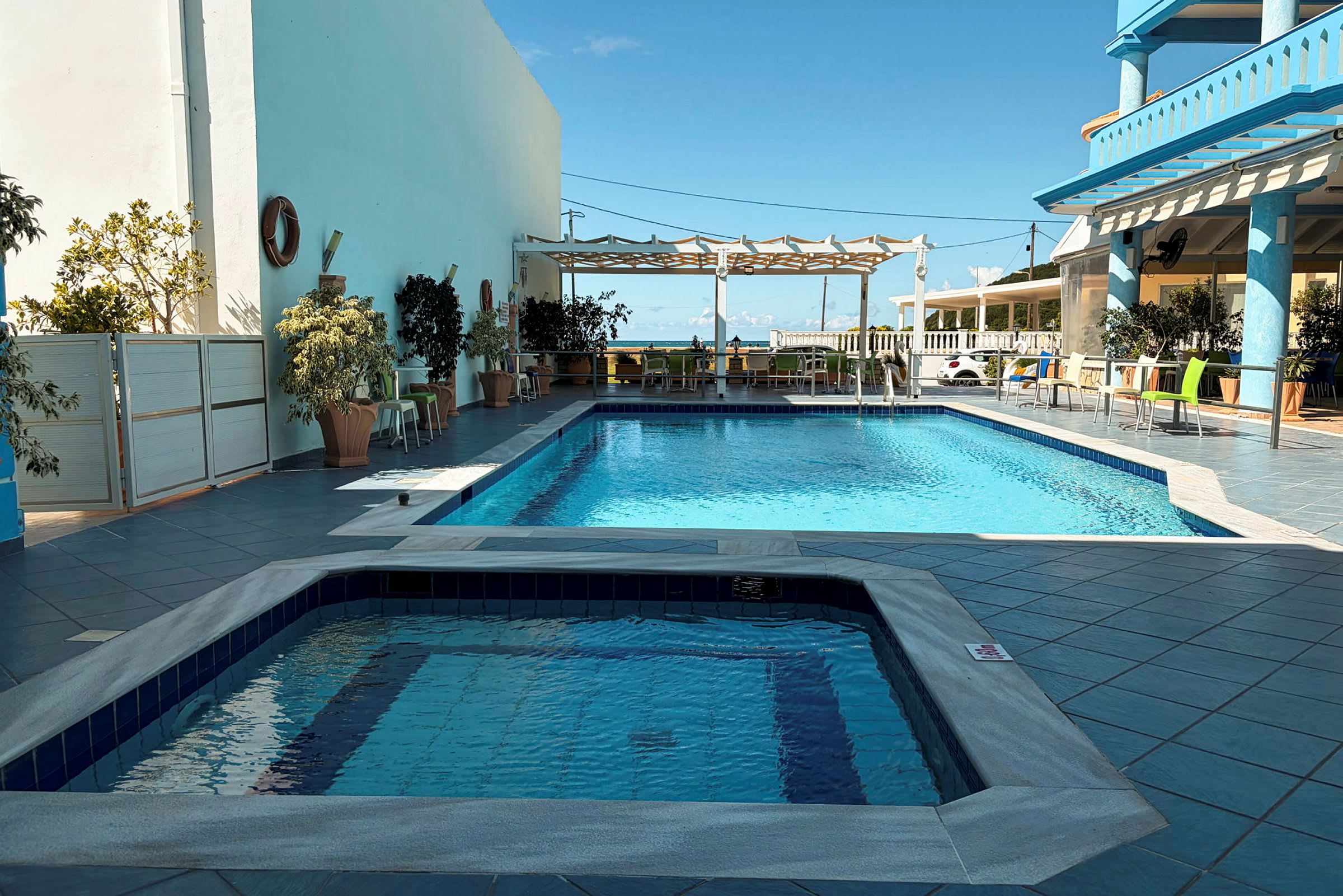 En utomhuspool med en separat jacuzzi i förgrunden, omgiven av bord, stolar och planteringar. (AI-genererad text)