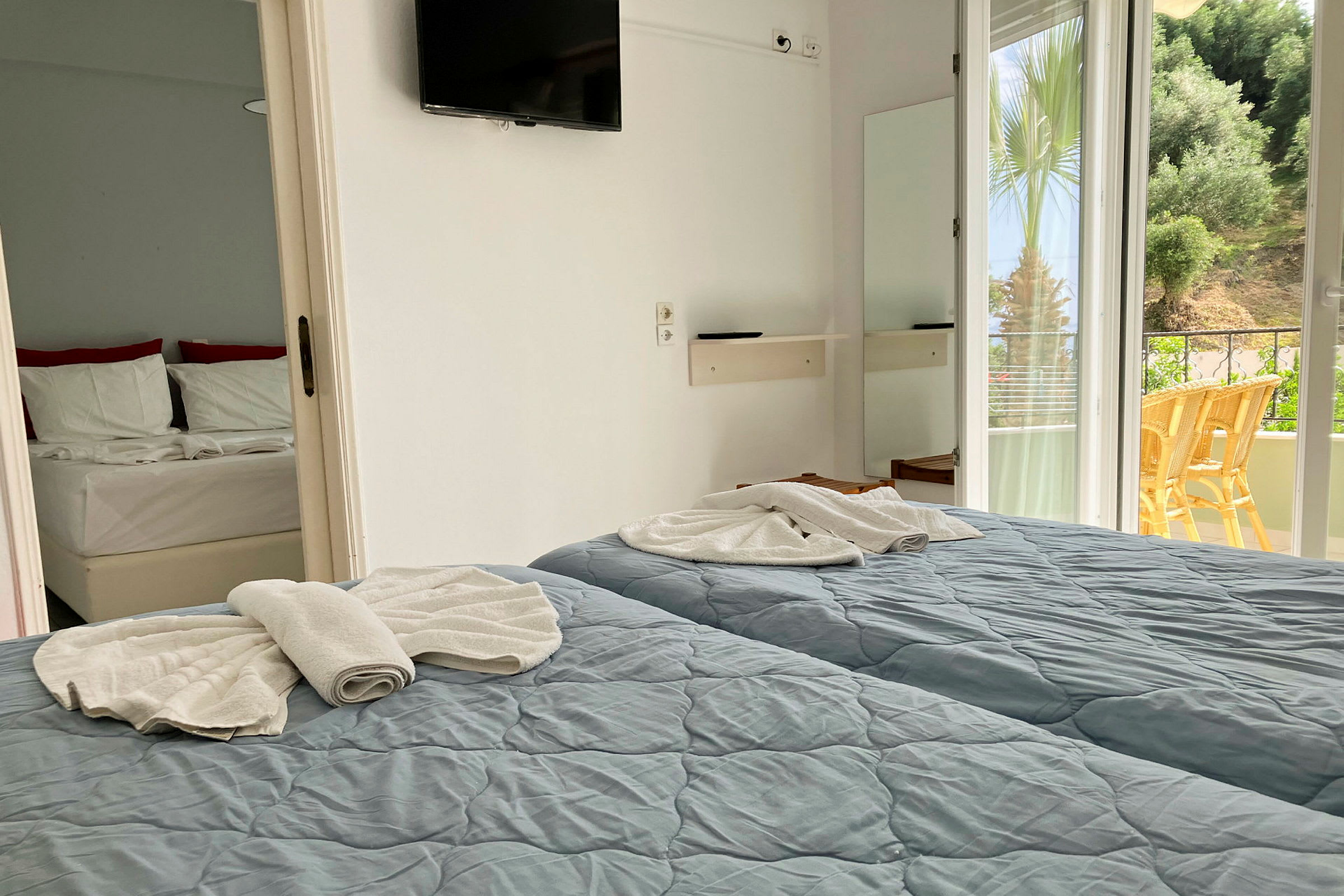 Ett hotellrum med två enkelsängar, en balkong med utsikt över grönska och en dörr till en annan del av rummet synlig. (AI-genererad text)