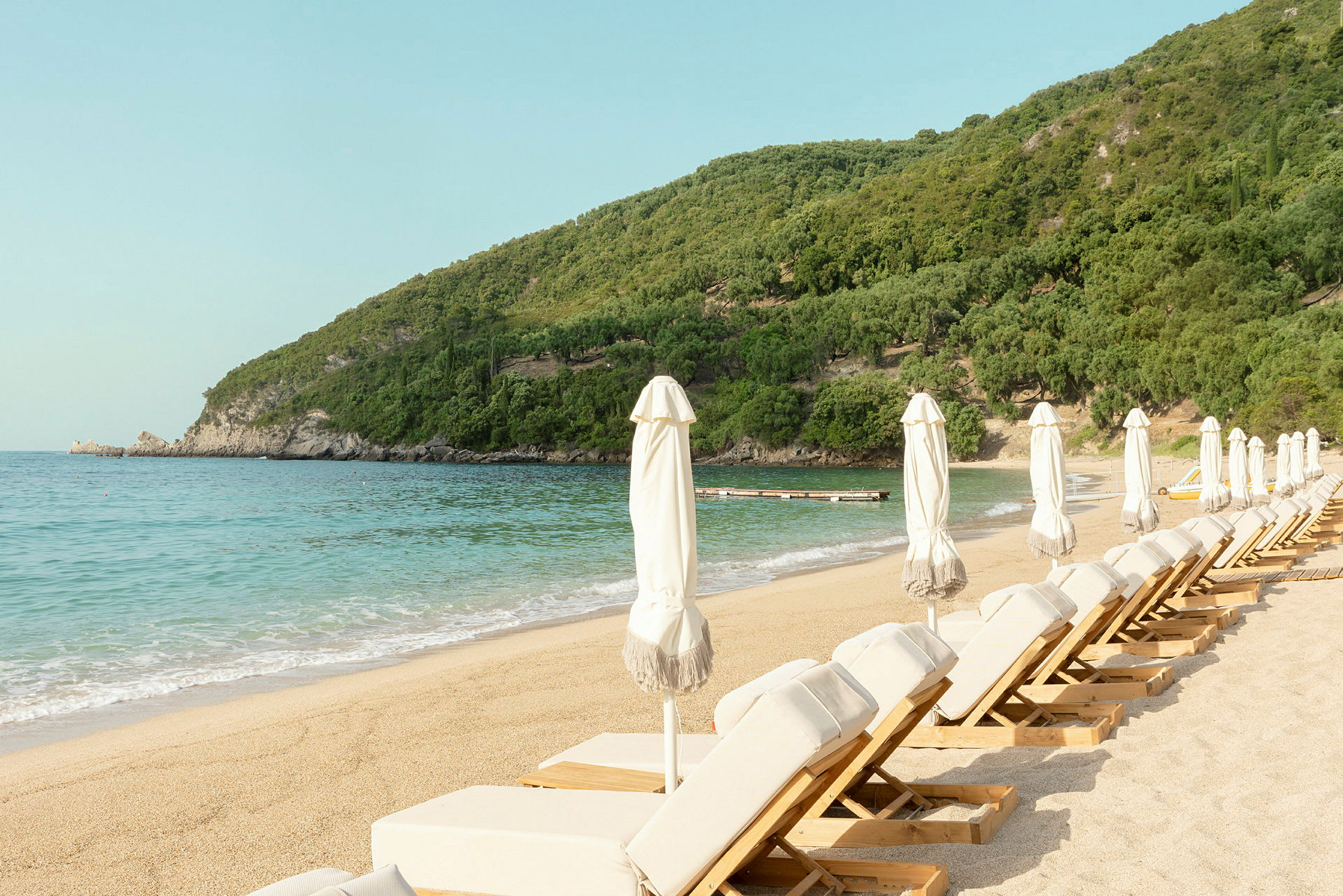 Solstolar och parasoller vid en sandstrand med hav och grönskande berg i bakgrunden. (AI-genererad text)