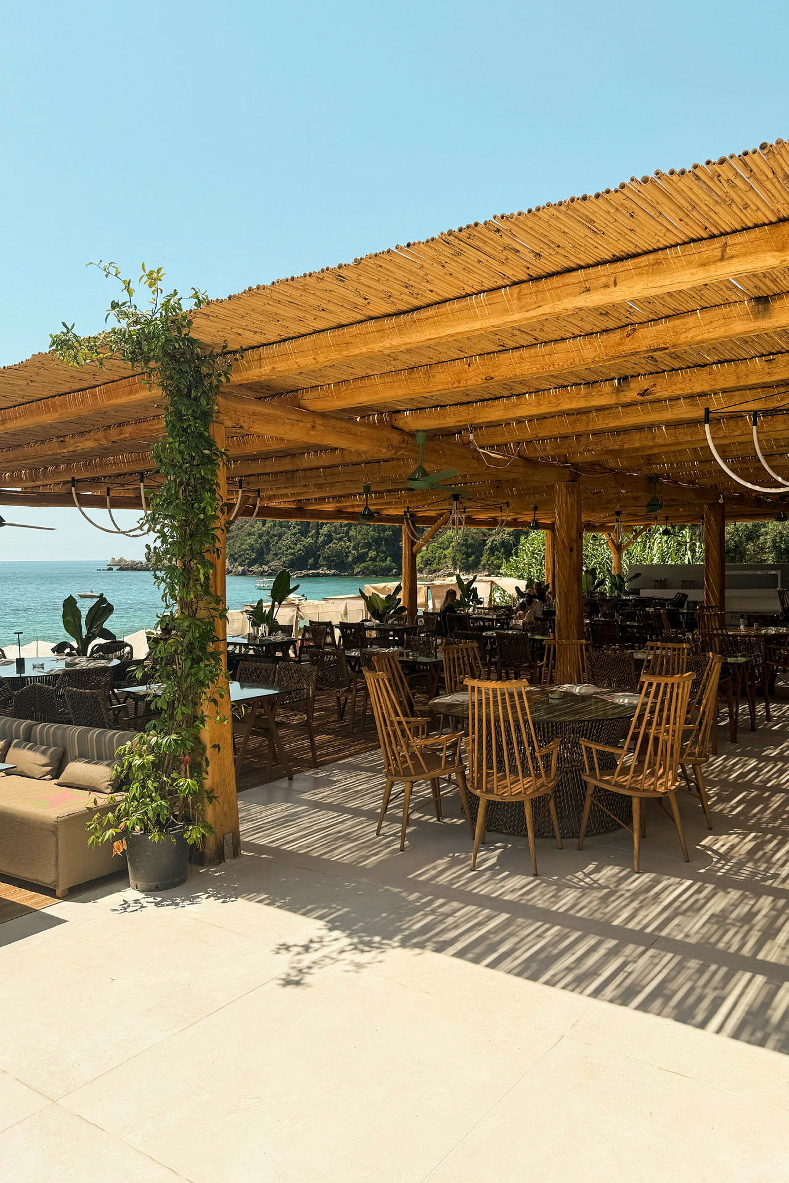 Utomhusservering med träpergola, bord och stolar samt utsikt mot havet. (AI-genererad text)