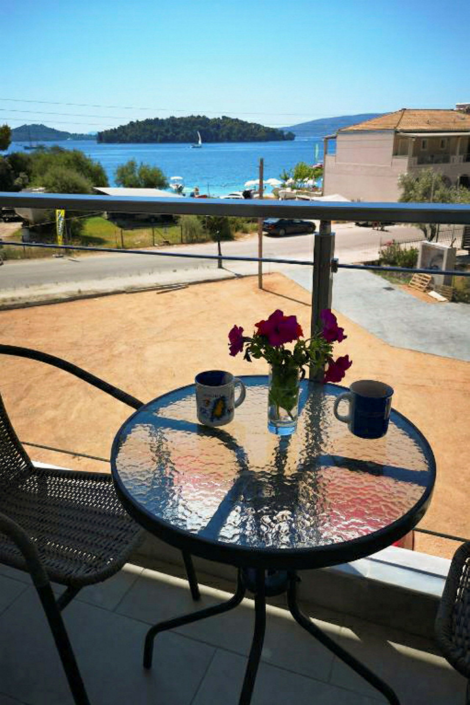 En balkong med ett glasbord, två koppar och en vas med rosa blommor, med utsikt mot havet och en liten ö. (AI-genererad text)