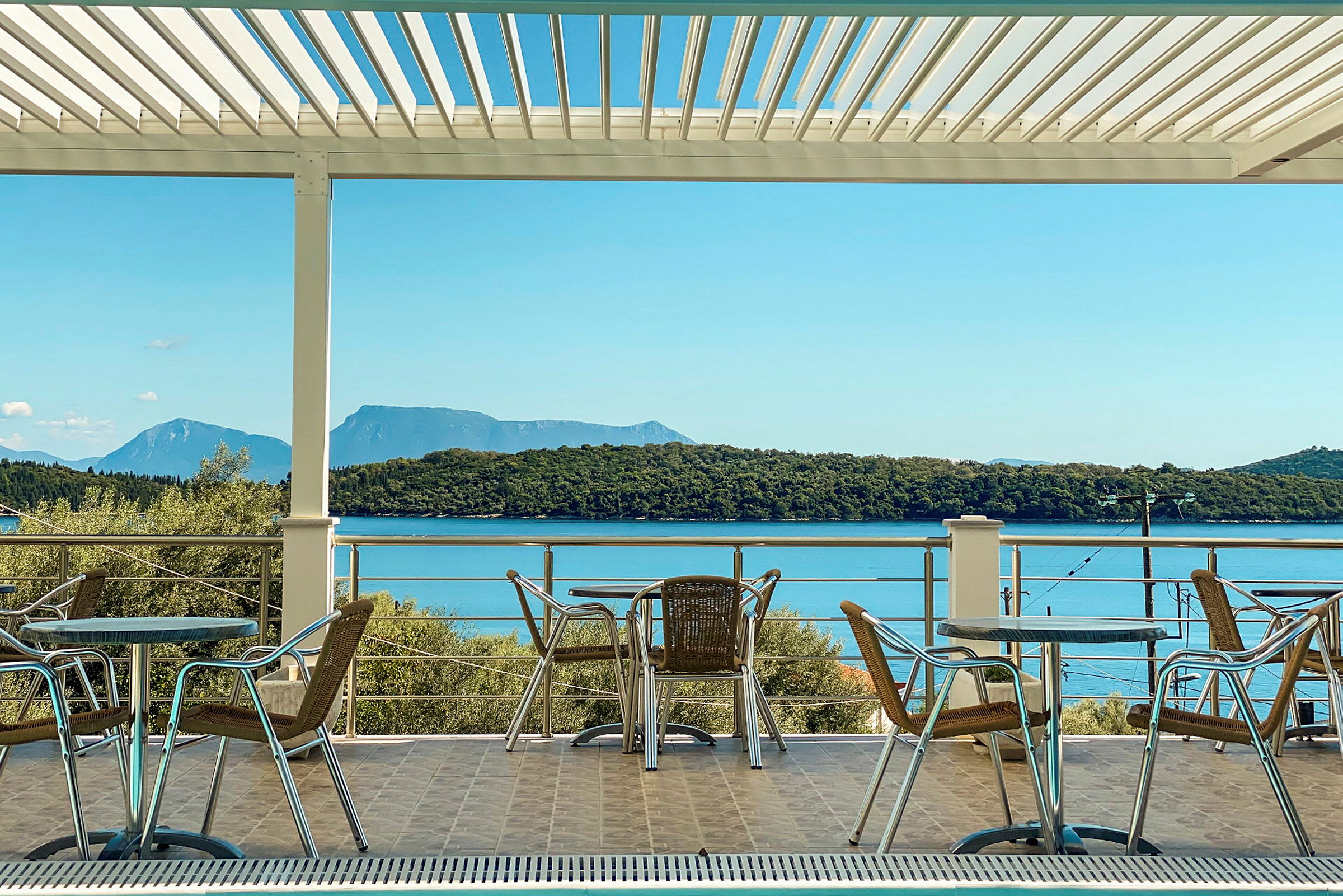 Utemöbler med bord och stolar på en terrass med utsikt över havet och skogbeklädda kullar. (AI-genererad text)