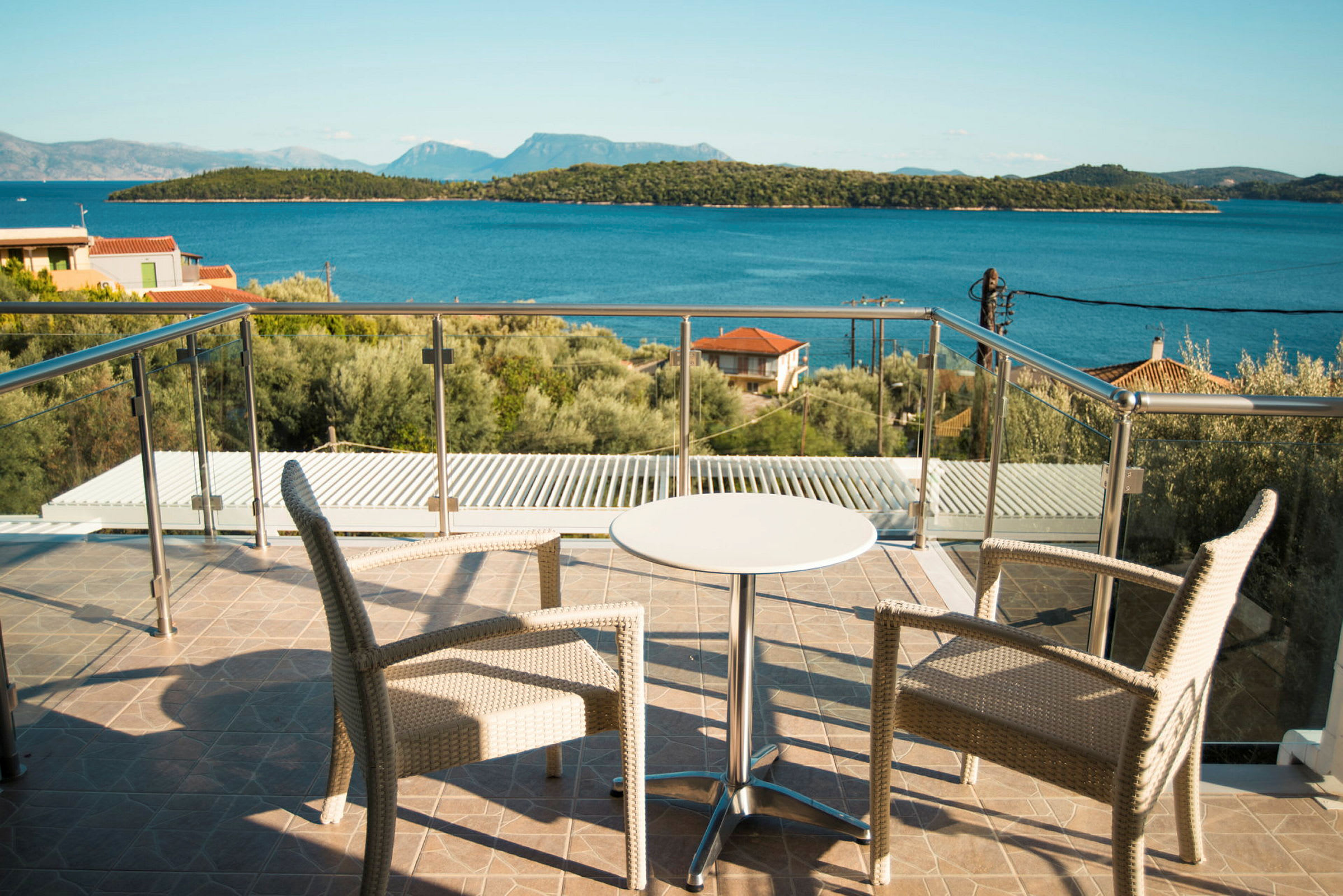 Två stolar och ett mindre bord på en terrass med glasräcke, utsikt mot havet och skogklädda öar. (AI-genererad text)