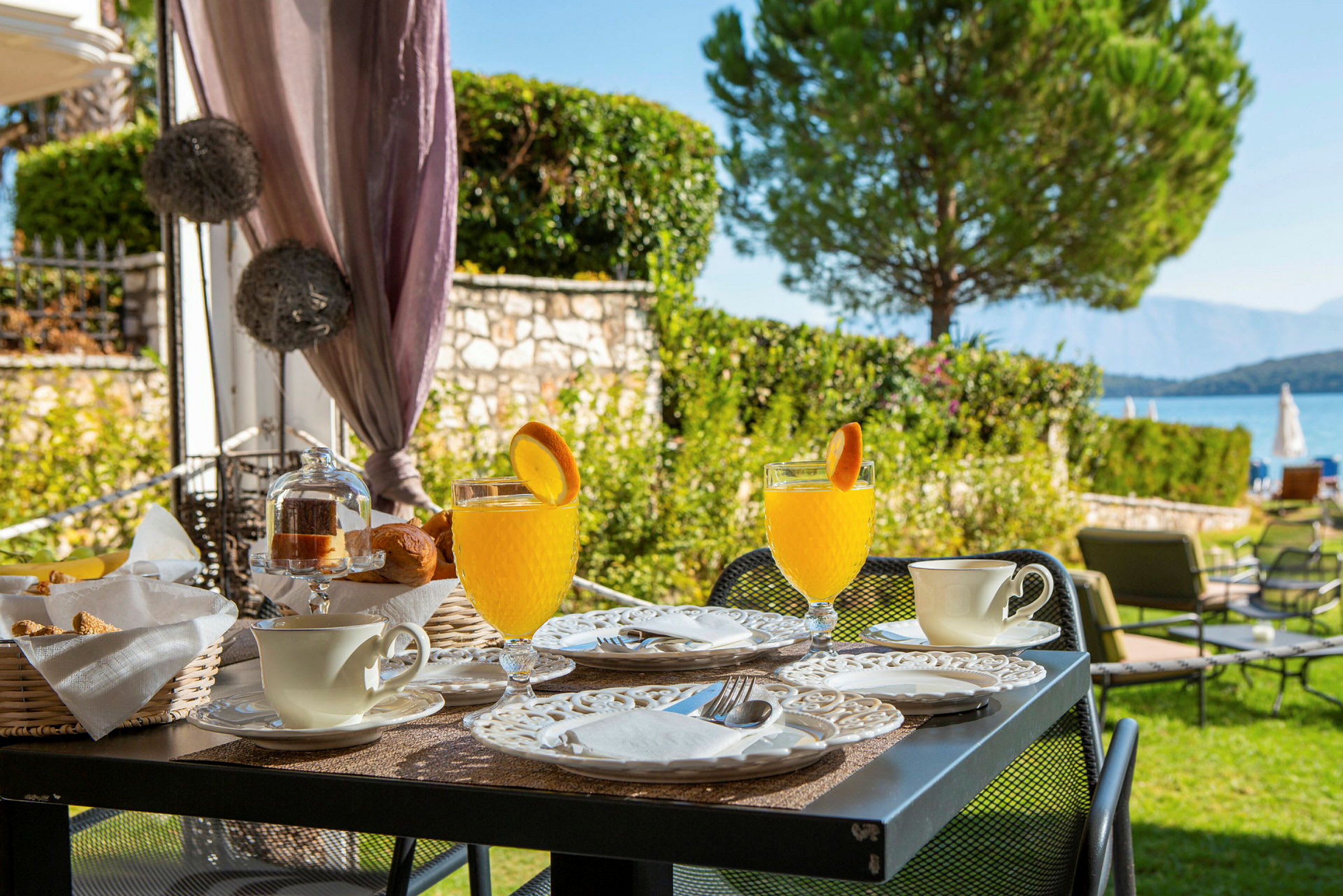 Ett bord med frukostservering, inklusive glas med juice dekorerade med apelsinskivor, koppar, och tallrikar, vid en trädgård nära havet. (AI-genererad text)