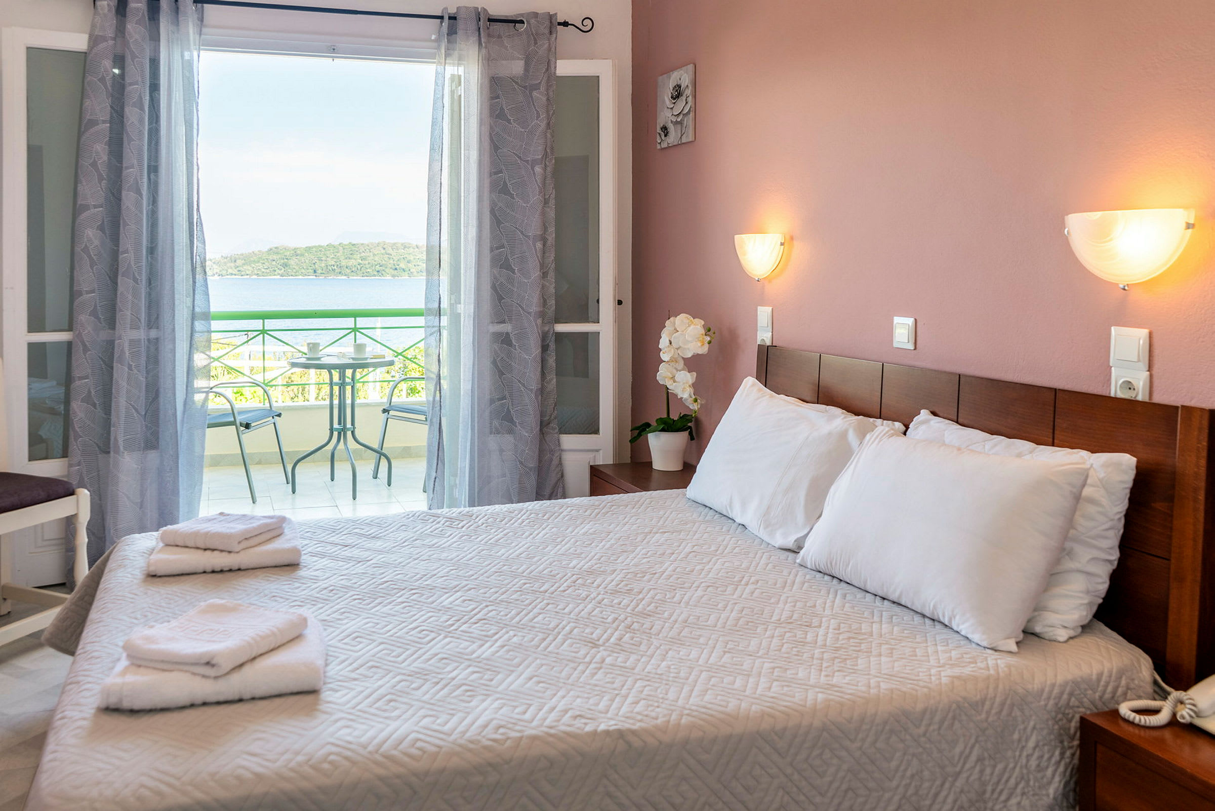 Ett hotellrum med en dubbelsäng, handdukar på sängen och en balkong i bakgrunden med utsikt mot havet. (AI-genererad text)