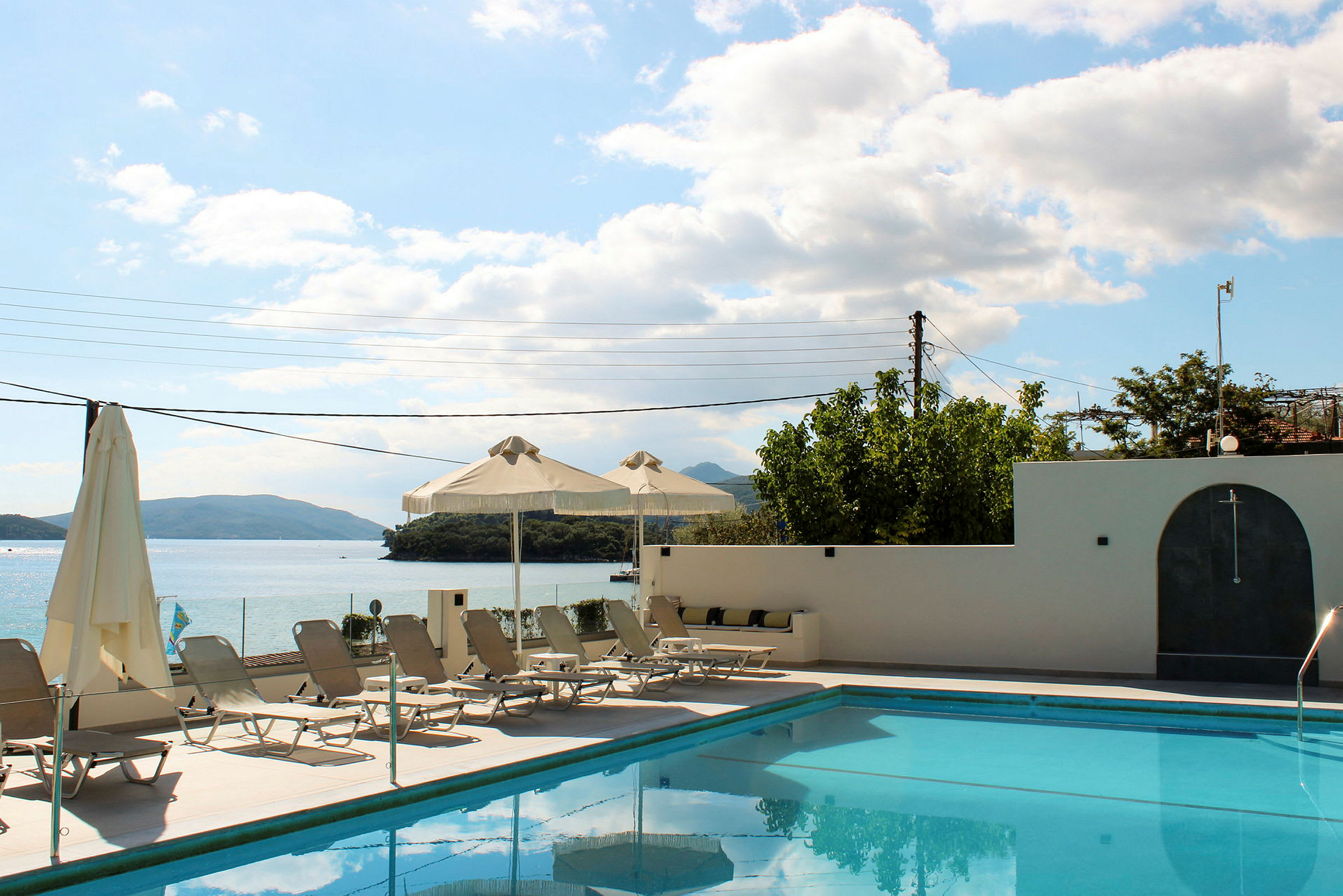 En swimmingpool vid en terrass med solstolar och parasoller, med utsikt över havet och berg i bakgrunden. (AI-genererad text)