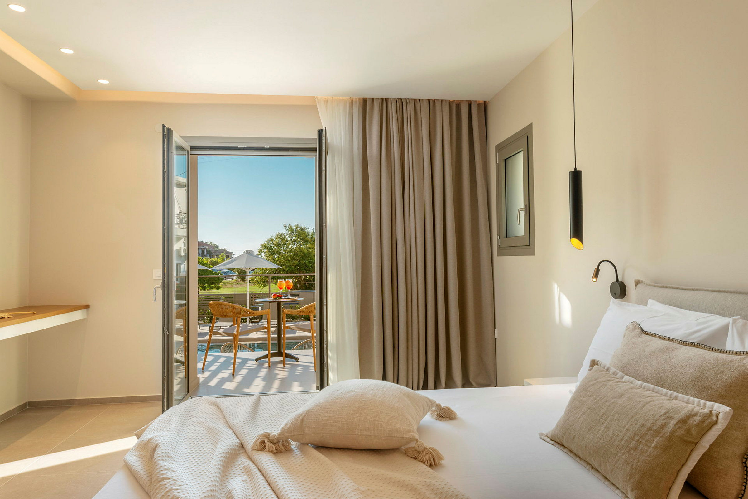 Ett hotellrum med en säng, en öppen dörr mot en balkong som har en sittgrupp och utsikt över grönska. (AI-genererad text)