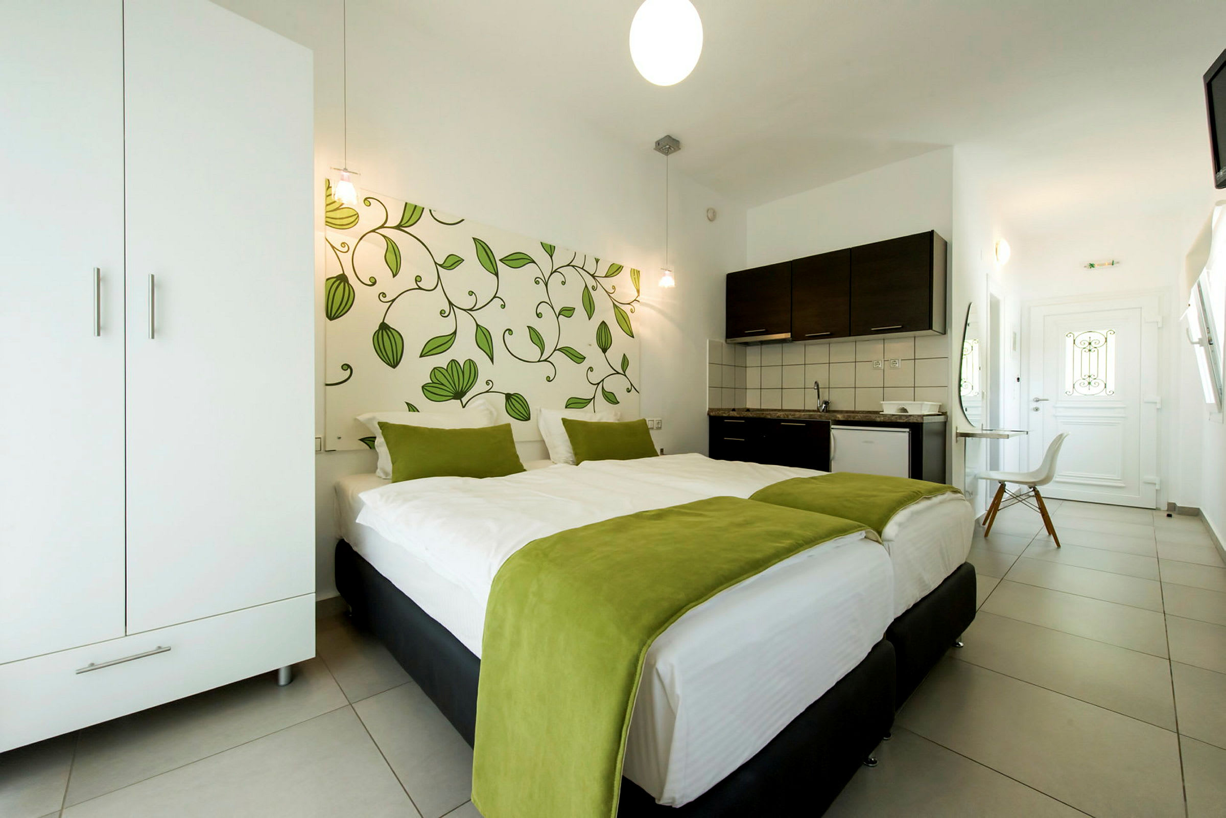 Ett hotellrum med en dubbelsäng täckt med vit sängkläder och gröna filtar, en garderob, ett pentry och en dörr i bakgrunden. (AI-genererad text)