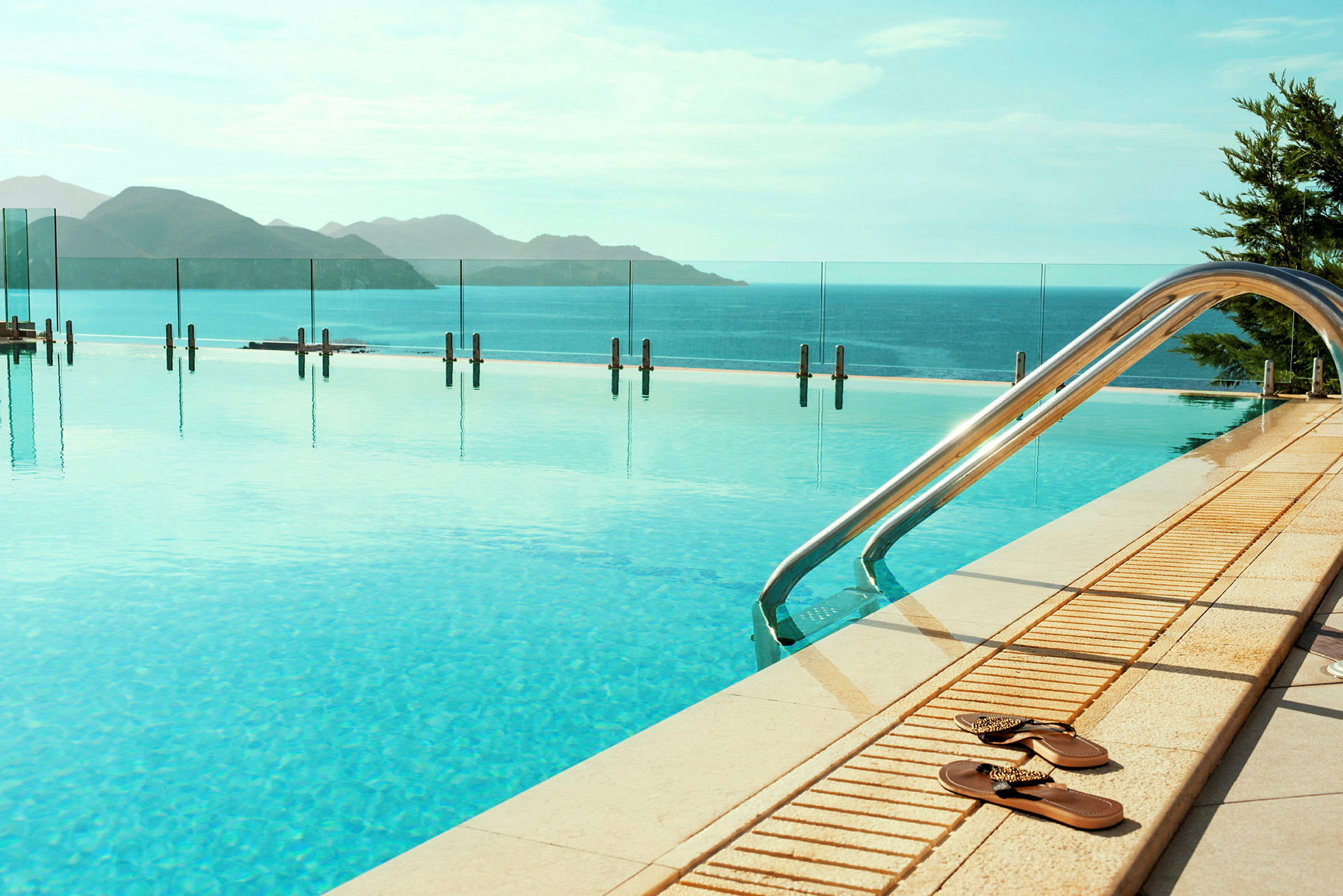 En utomhus infinitypool med utsikt över havet och berg i bakgrunden, samt ett par flip-flops vid kanten. (AI-genererad text)