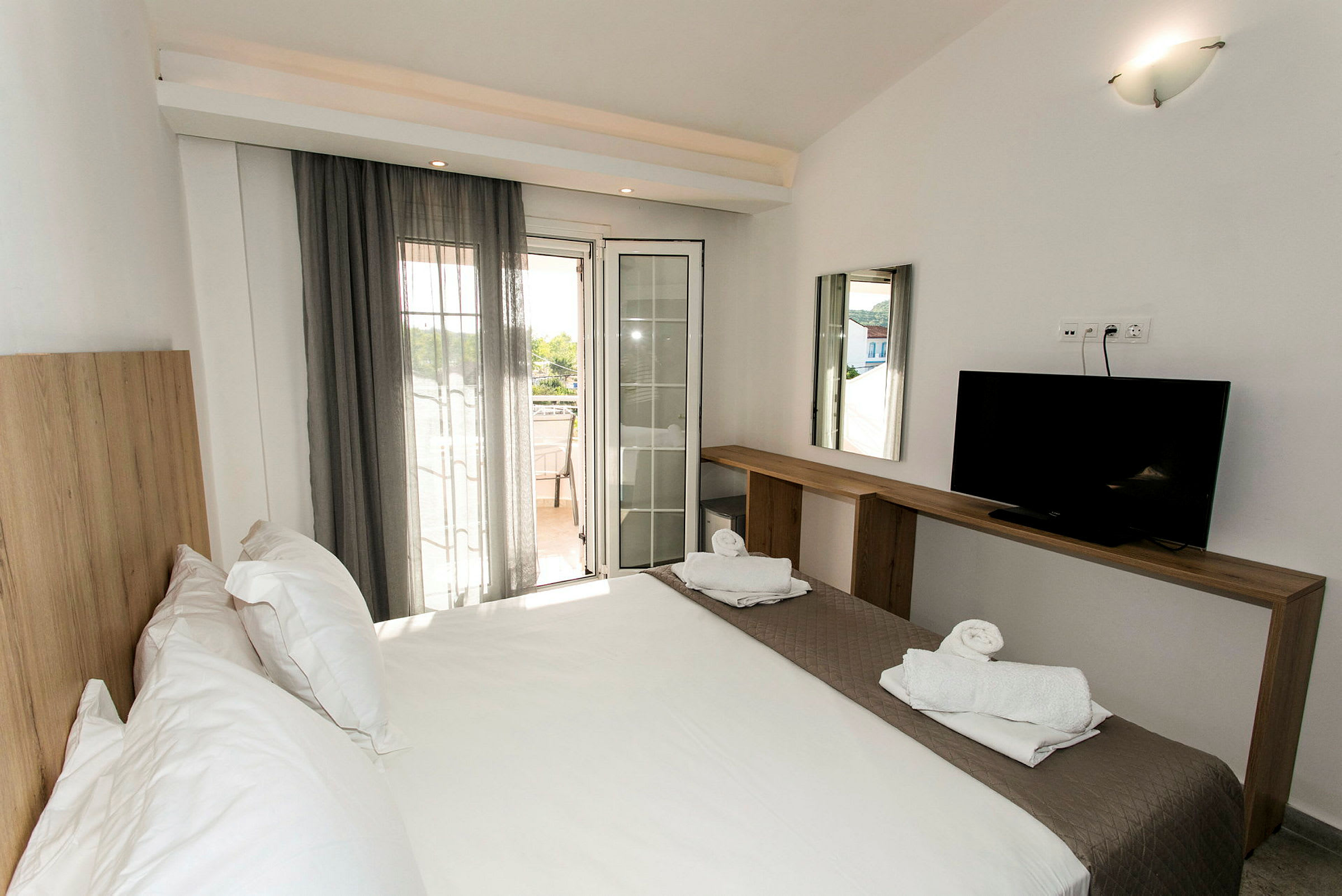 Ett hotellrum med en dubbelsäng, en TV på en träbänk, och en balkong med utsikt. (AI-genererad text)