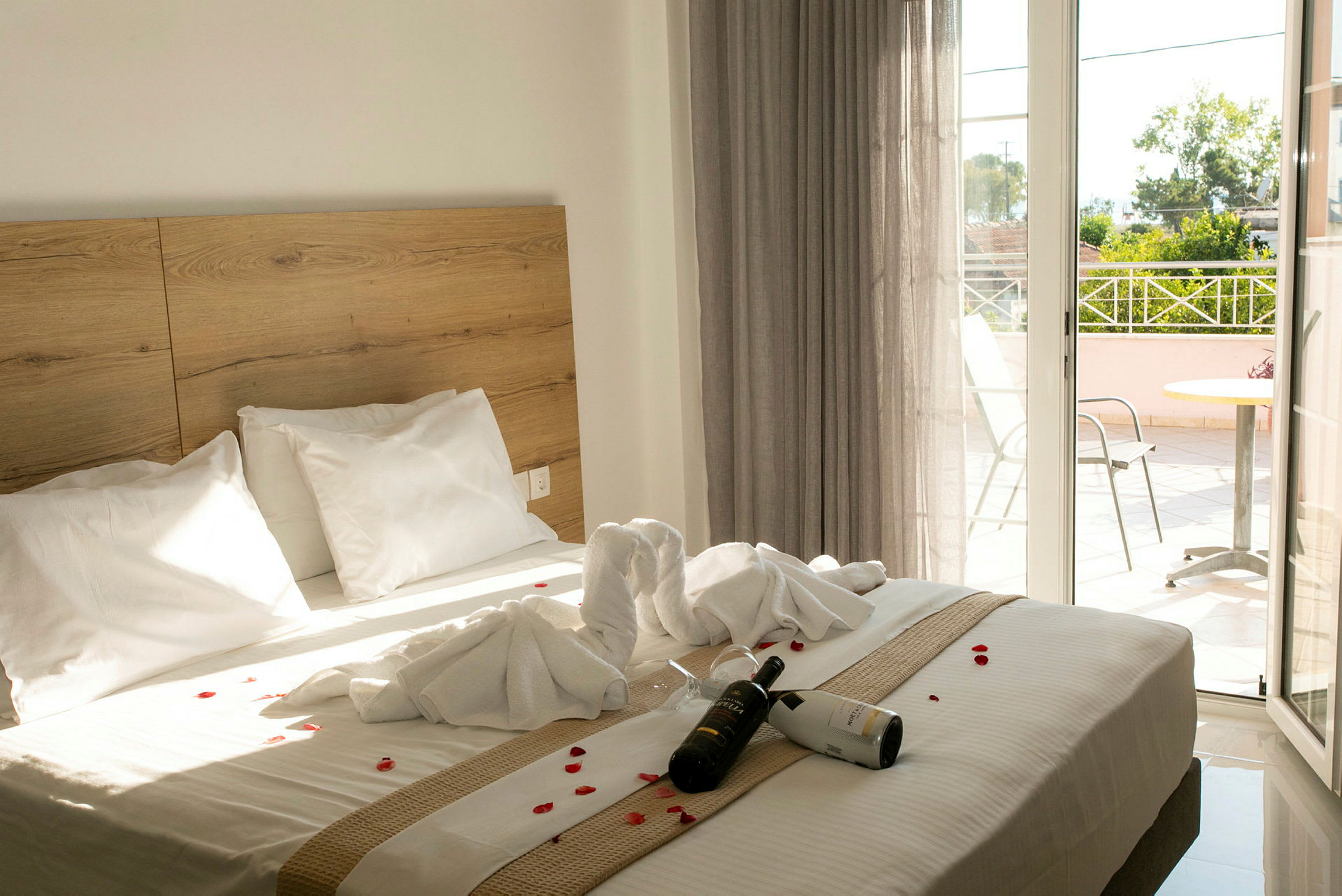 Hotellrum med en dubbelsäng dekorerad med handdukar och blomblad. En flaska vin och glas ligger på sängen. En balkong är synlig i bakgrunden. (AI-genererad text)