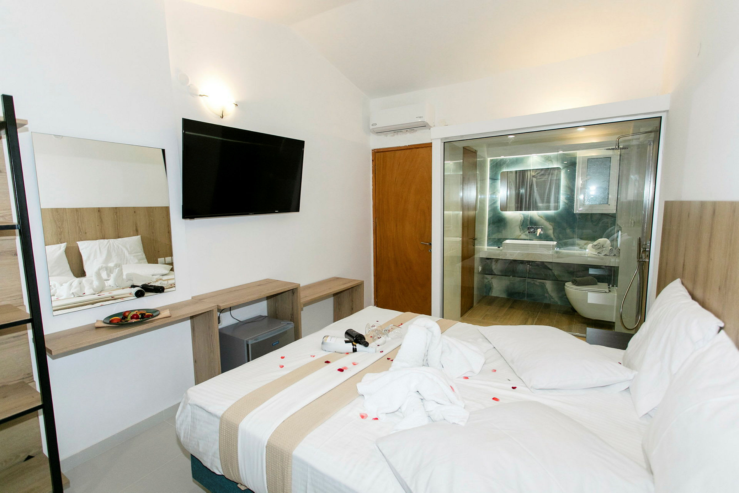Hotellrum med en stor säng, ett öppet badrum med glasvägg, och en uppsättning möbler inklusive en TV. (AI-genererad text)