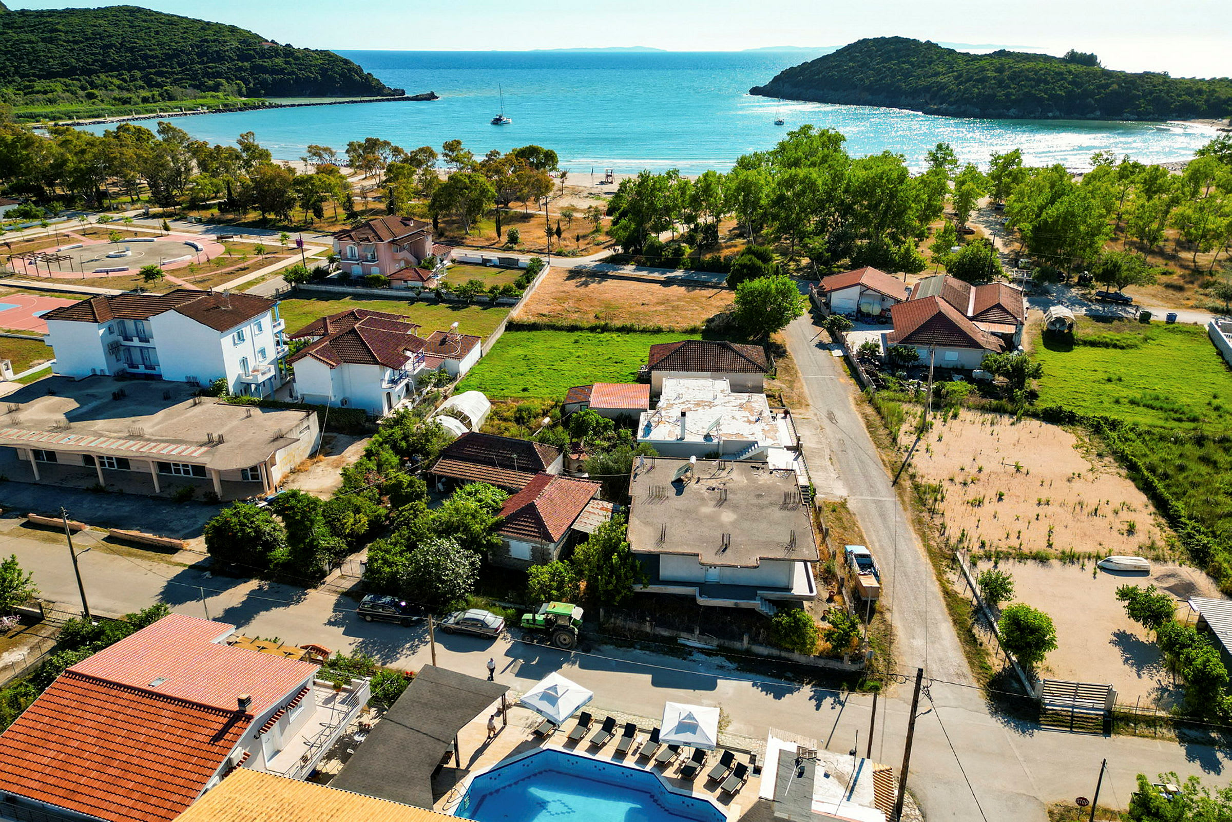 Flygfoto av ett kustområde med byggnader, väg och grönytor nära havet med båtar. (AI-genererad text)