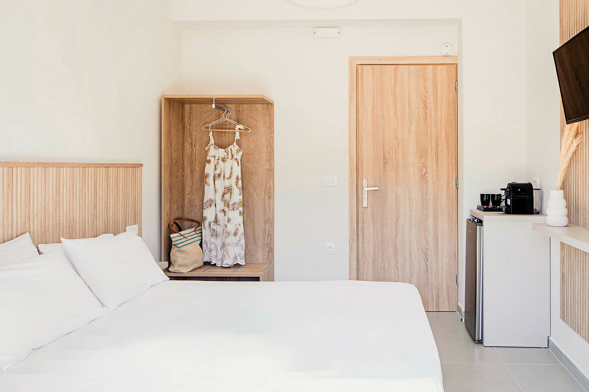 Ett hotellrum med en vit säng, en garderob med en klänning och väska, en dörr och ett litet köksområde med en kaffemaskin. (AI-genererad text)