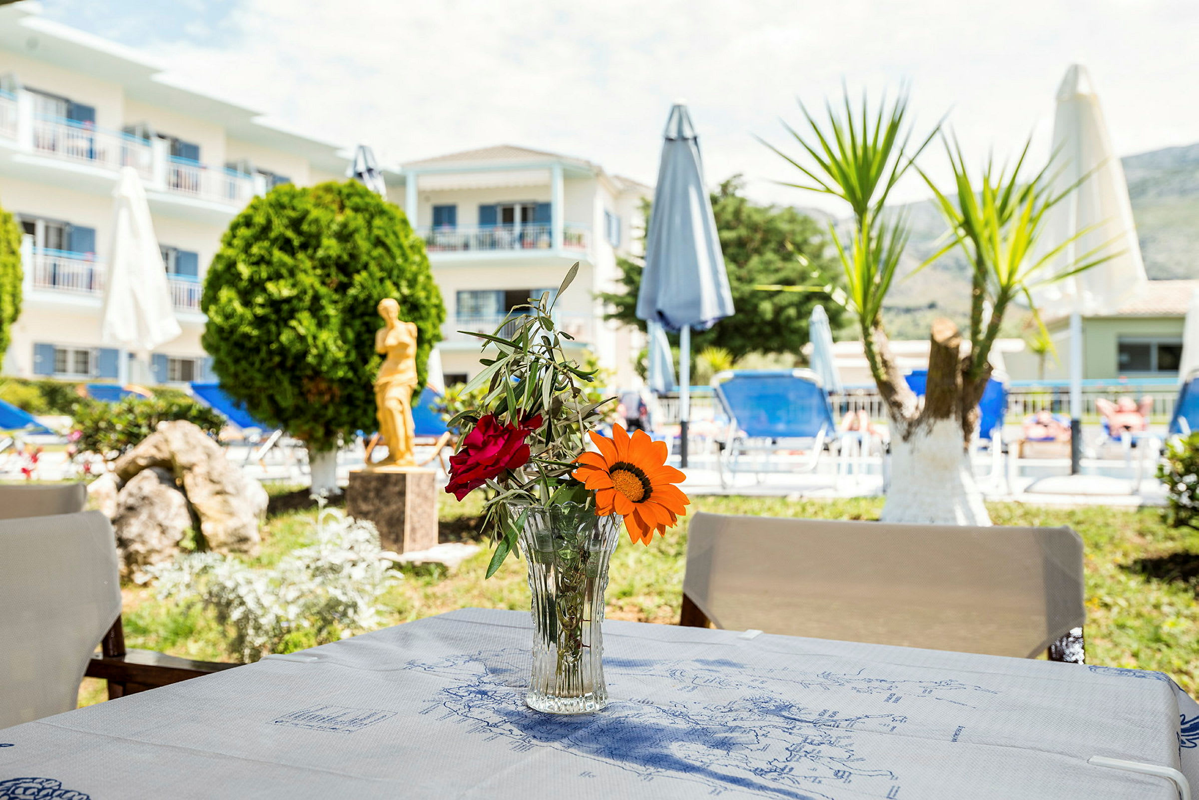 Bild på ett bord med en vas innehållandes blommor, i bakgrunden syns en byggnad, parasoller och en poolområde. (AI-genererad text)