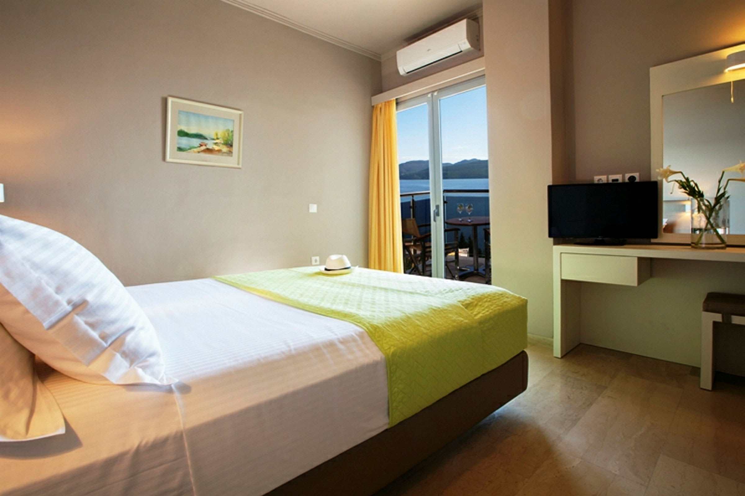 Ett hotellrum med en säng, skrivbord, spegel och utsikt över en balkong med hav i bakgrunden. (AI-genererad text)
