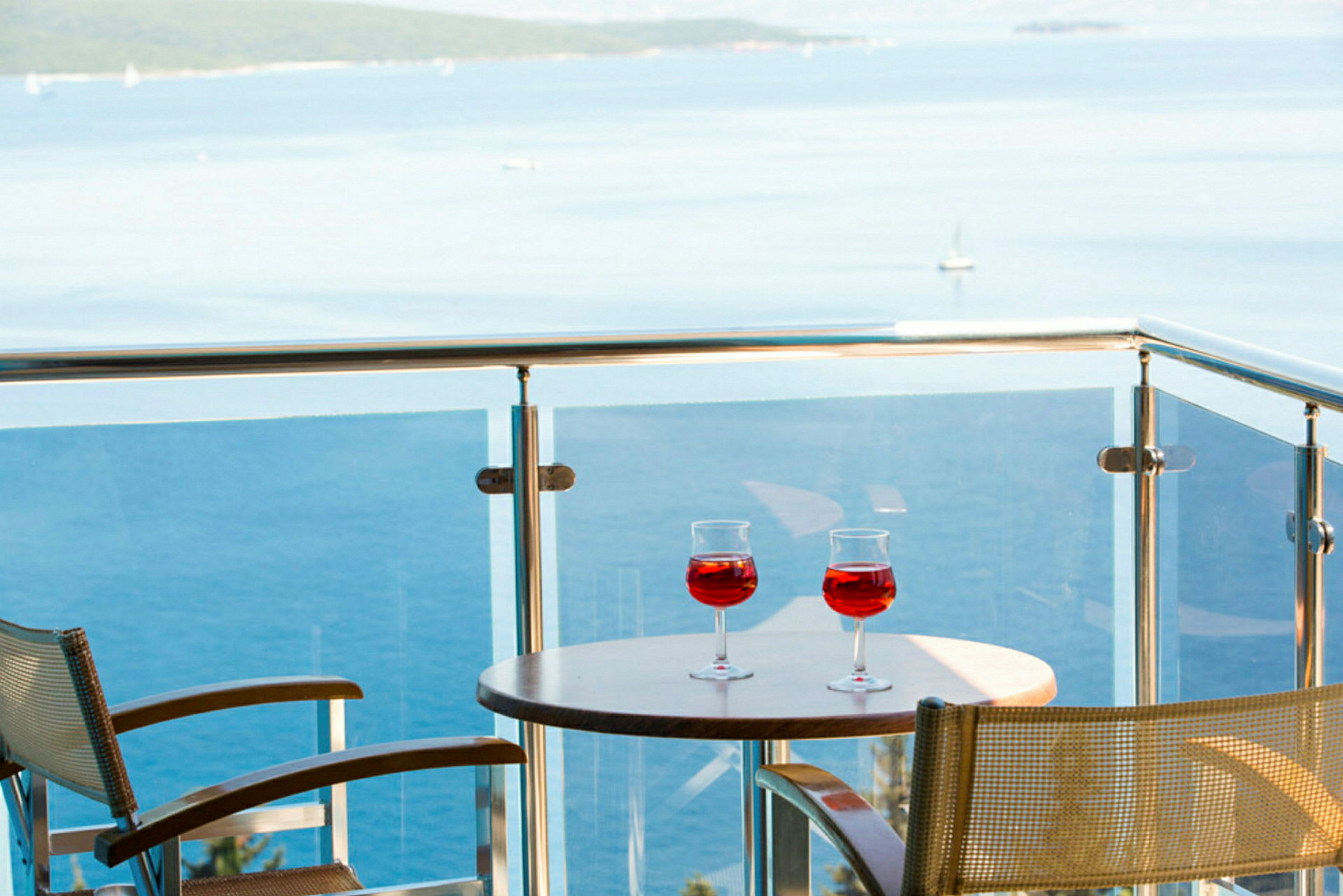 Två glas med röd dryck på ett bord på en balkong med utsikt över havet. (AI-genererad text)