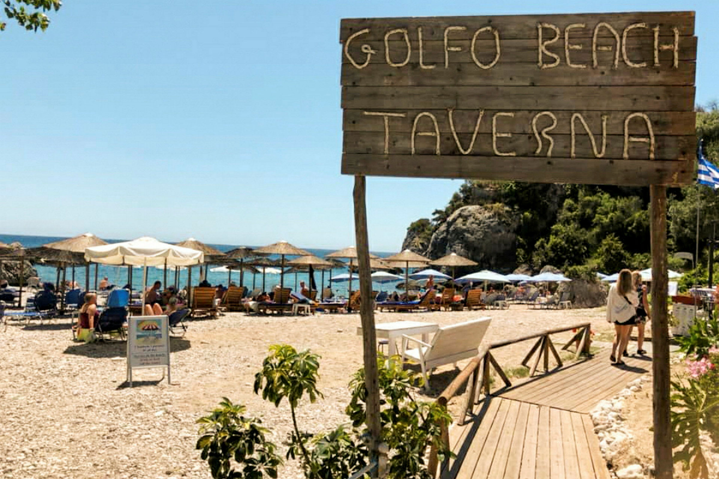 Skylt med texten 'Golfo Beach Taverna' framför en strand med solparasoller och solstolar. (AI-genererad text)