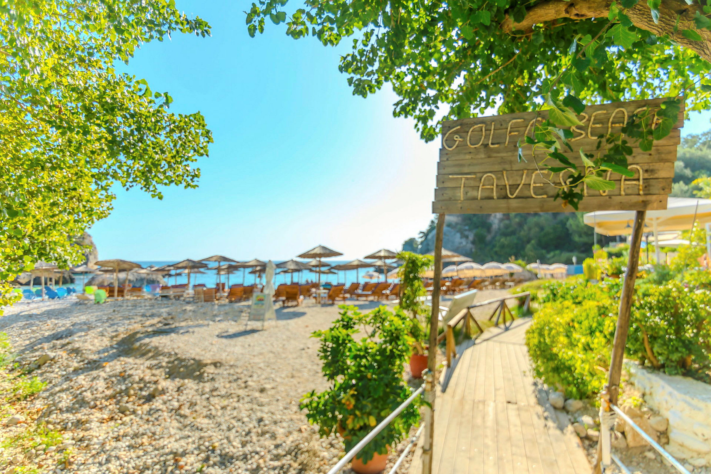 Skylt med texten 'Golf Beach Taverna' vid ingången till en strand med parasoller och solstolar. (AI-genererad text)