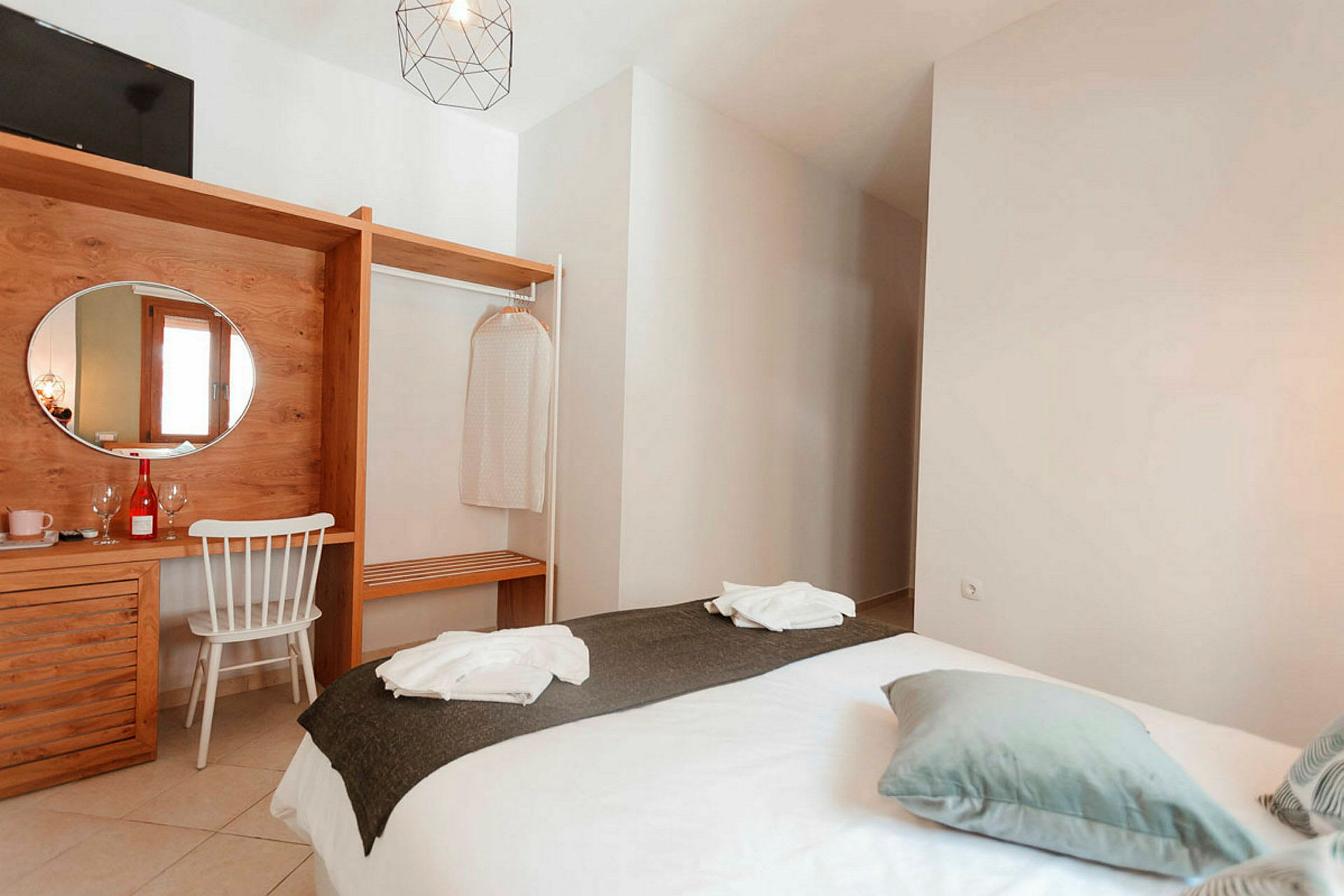 Ett hotellrum med en säng klädd i vita lakan och mörkgrå överkast, en trämöbel med en spegel och en stol, och detaljer som klädhängare och dekoration. (AI-genererad text)