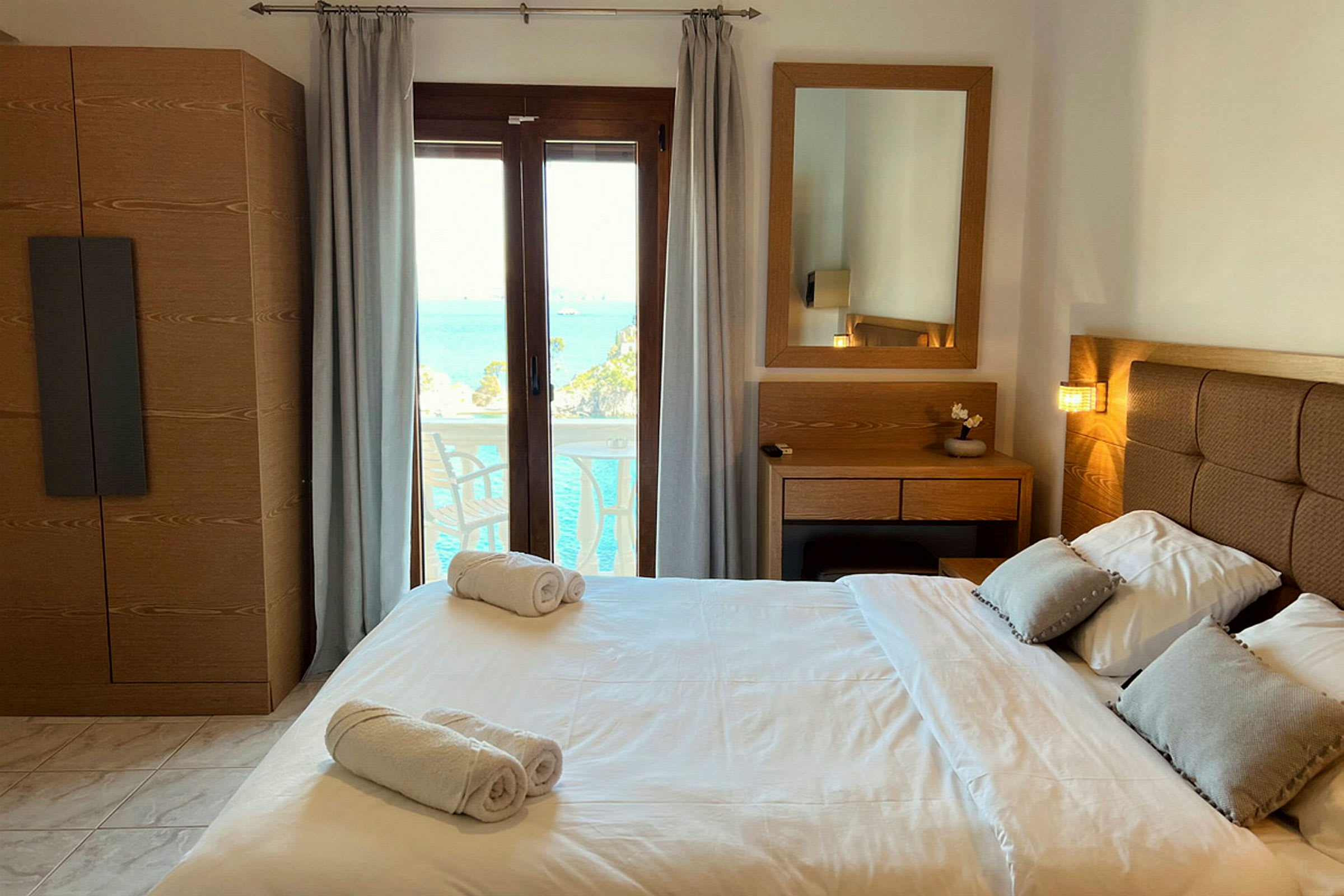 Ett hotellrum med en dubbelsäng, sängbord, spegel och utsikt över havet genom ett glasfönster. (AI-genererad text)