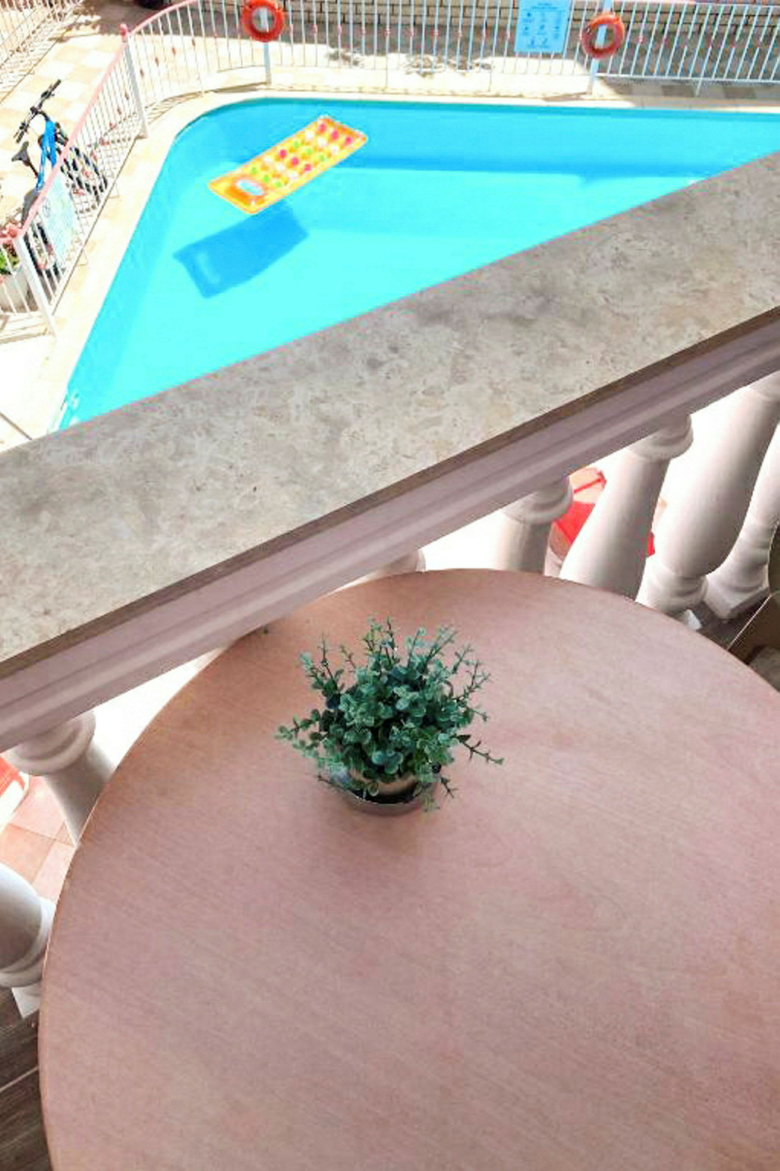 En utsikt från en balkong mot en pool med en badleksak och ett bord med en krukväxt. (AI-genererad text)