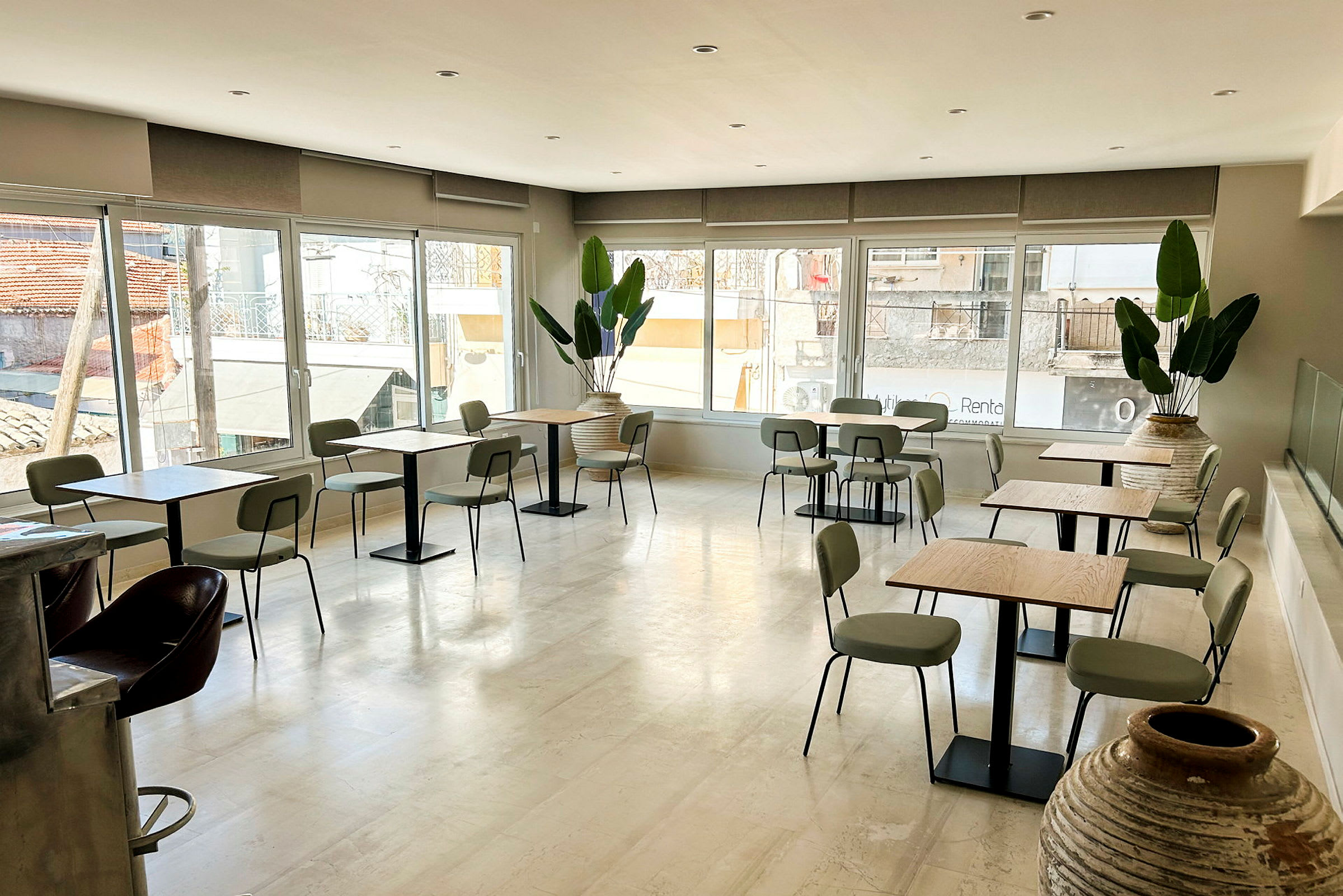 Ett ljust kafé med bord och stolar samt stora fönster som släpper in naturligt ljus. Flera krukväxter pryder rummet. (AI-genererad text)