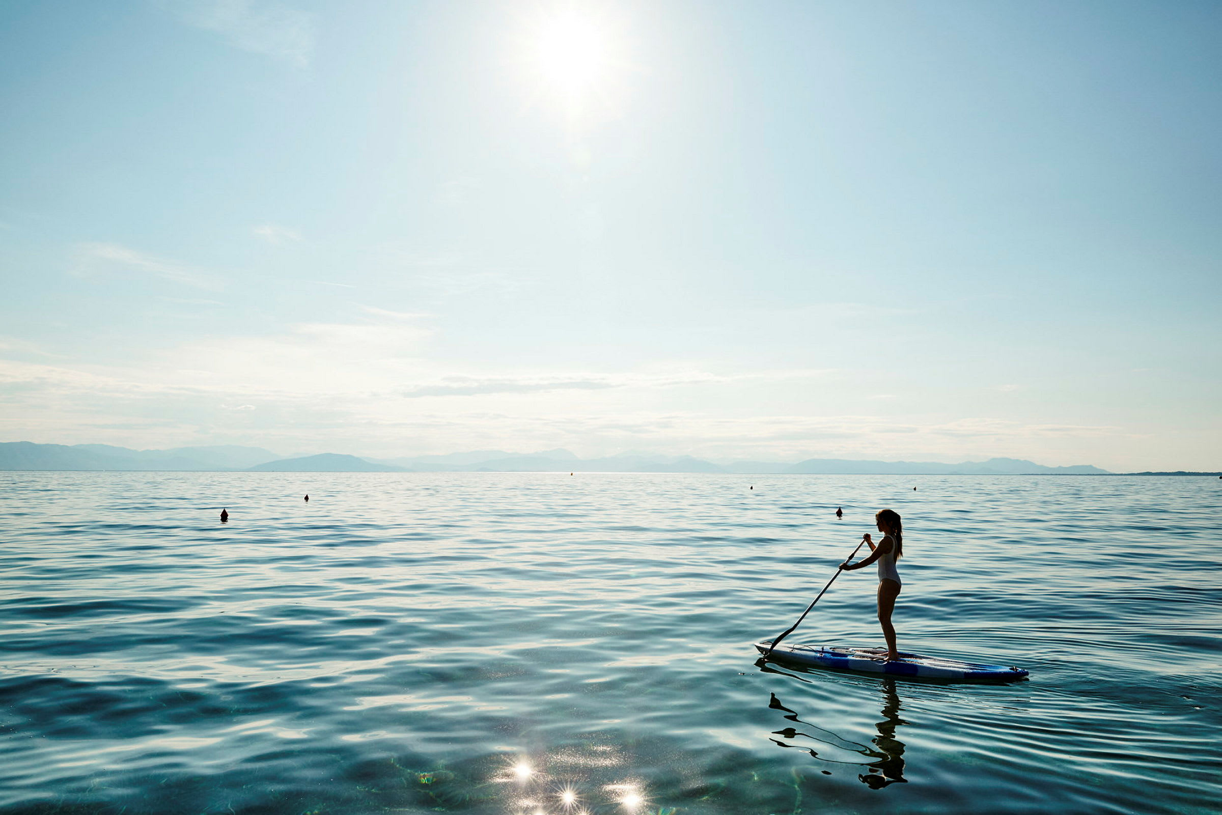 Kvinna som står på en paddleboard i en lugn sjö med solglitter och berg i bakgrunden. (AI-genererad text)