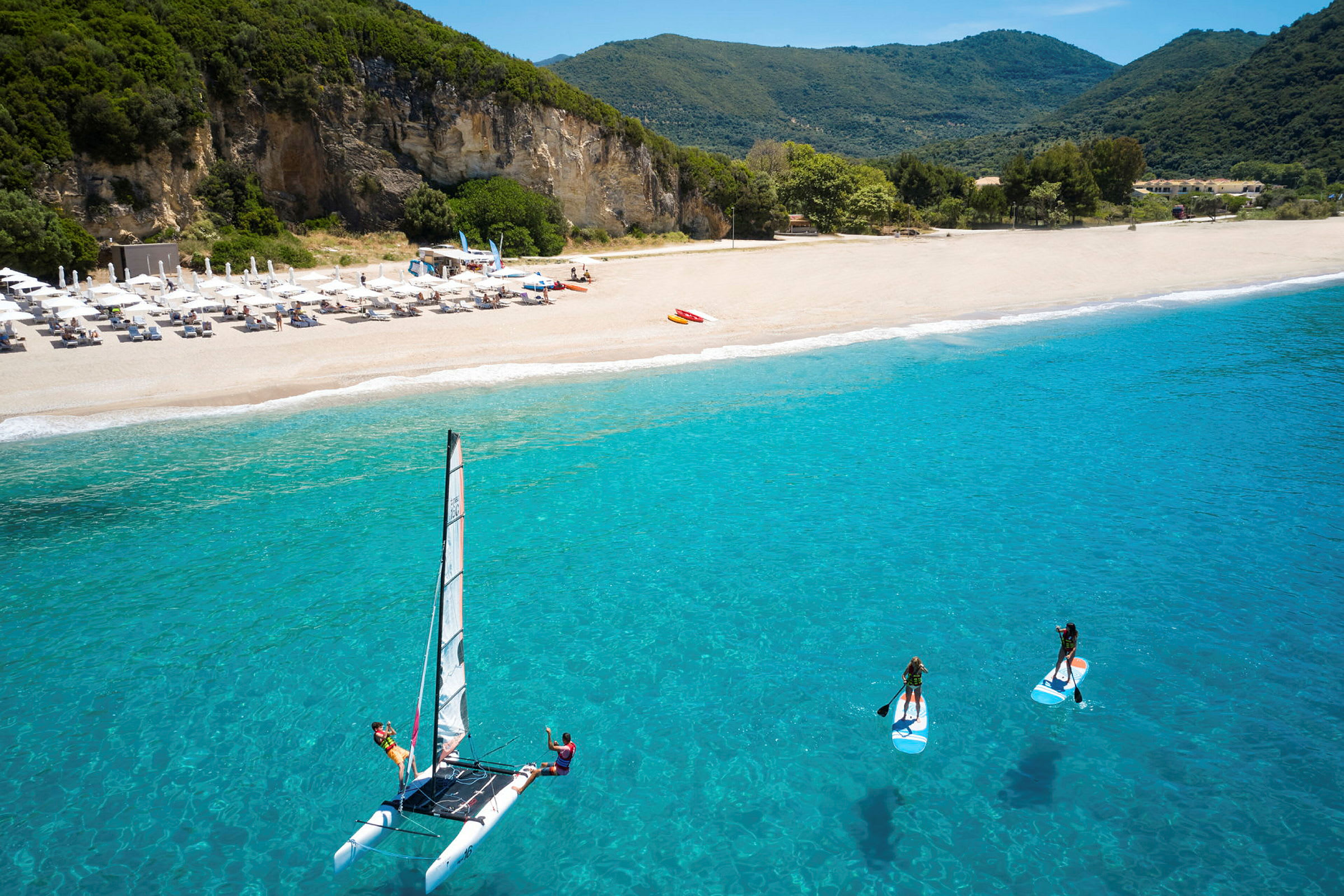 En strand med parasoller, klippor i bakgrunden och aktiviteter på vattnet, inklusive segling och paddleboarding. (AI-genererad text)