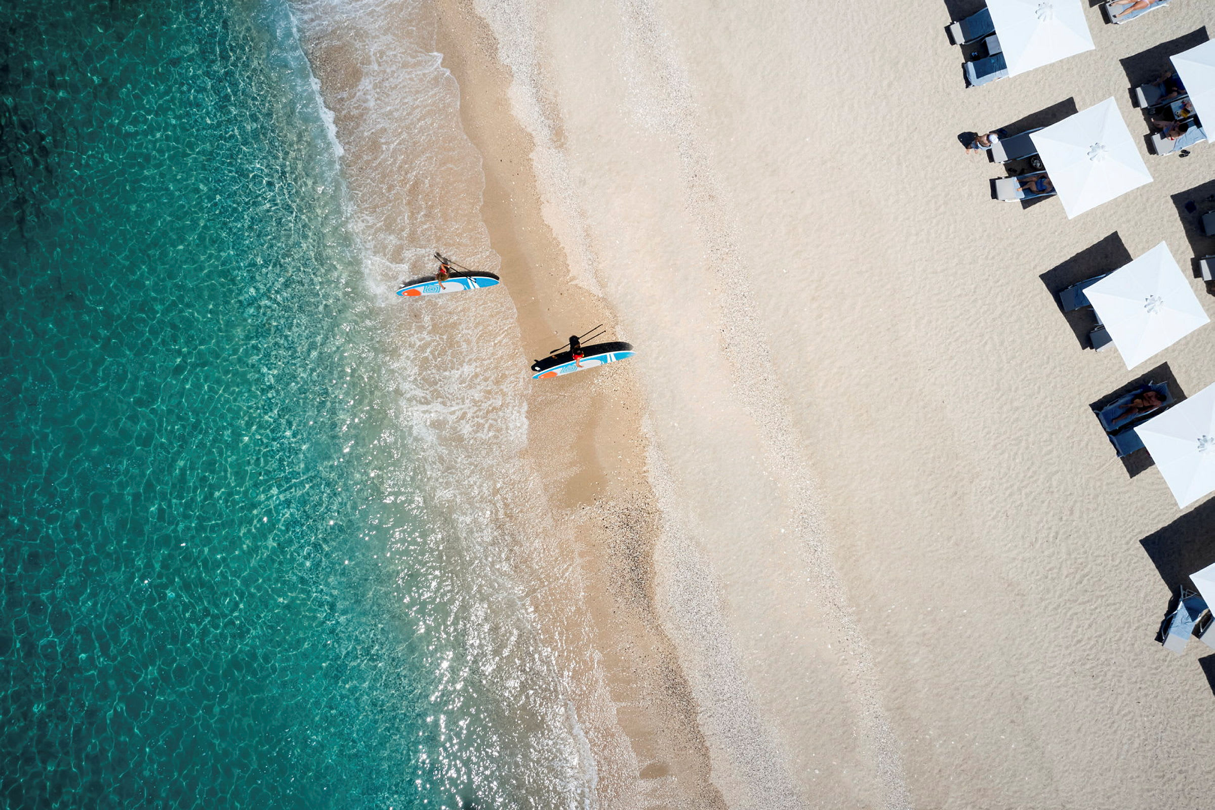 En strand med parasoller och personer som paddlar på surfbrädor nära vattenbrynet. (AI-genererad text)