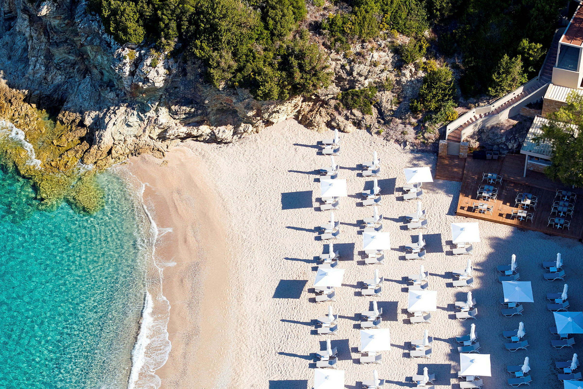 Flygbild på en strand med parasoller och solstolar nära klippor och klarblått hav. (AI-genererad text)