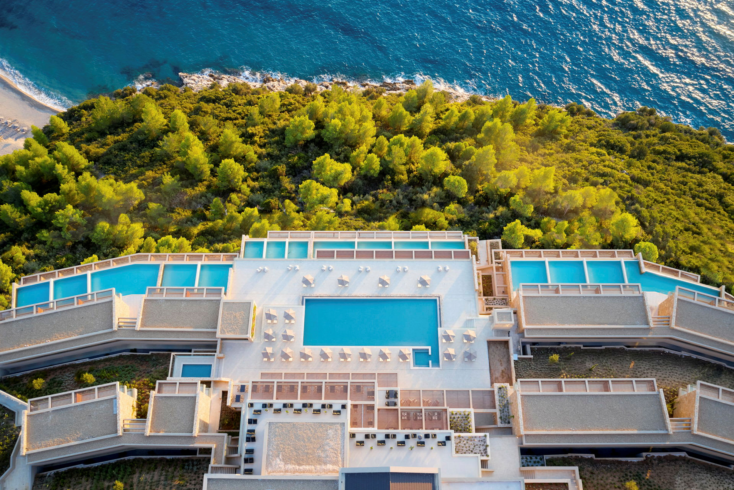 Flygbild av hotellområde med swimmingpooler omgiven av grönska nära havet. (AI-genererad text)