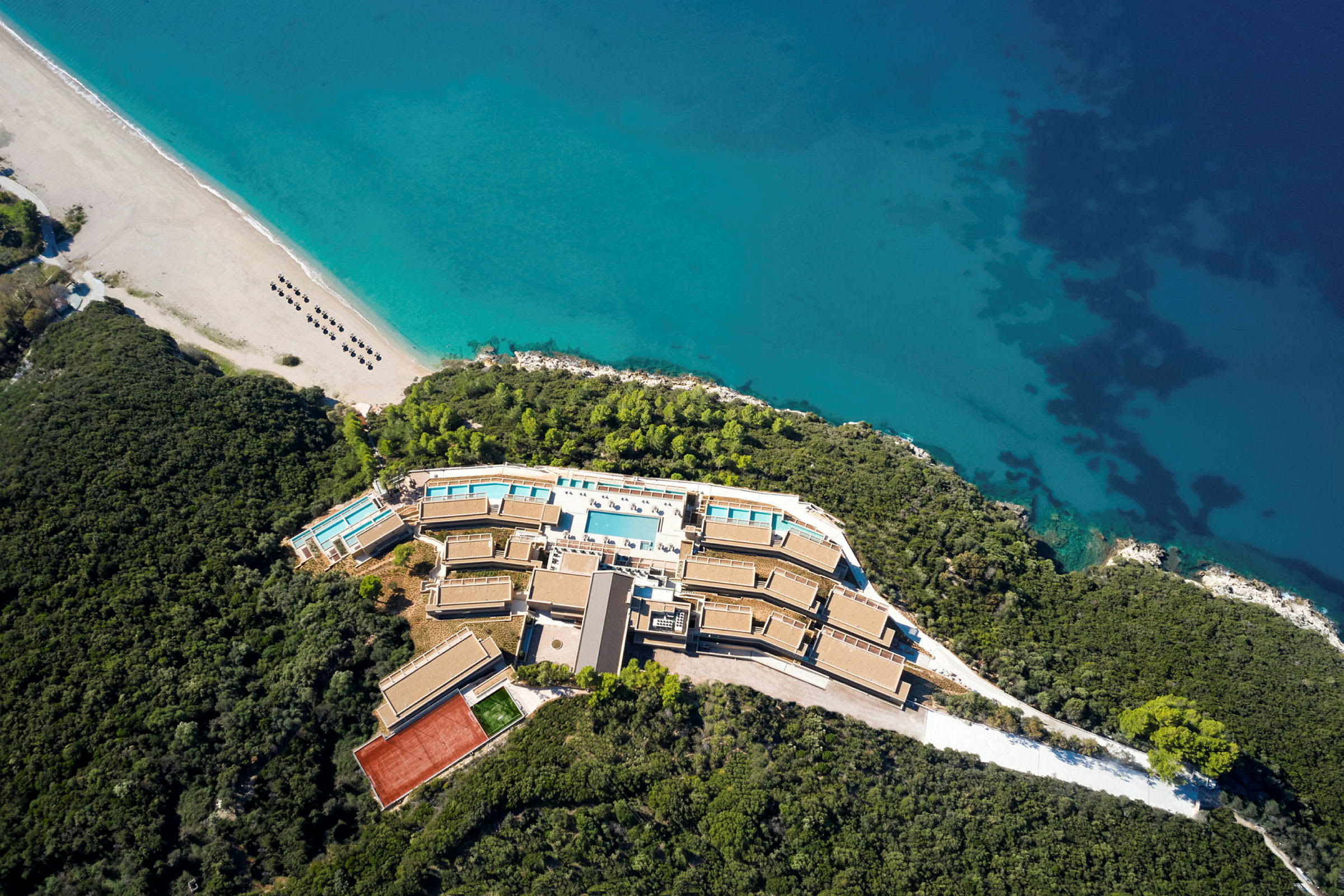 Flygfoto av en resort vid en strand med gröna omgivningar och blått hav. (AI-genererad text)