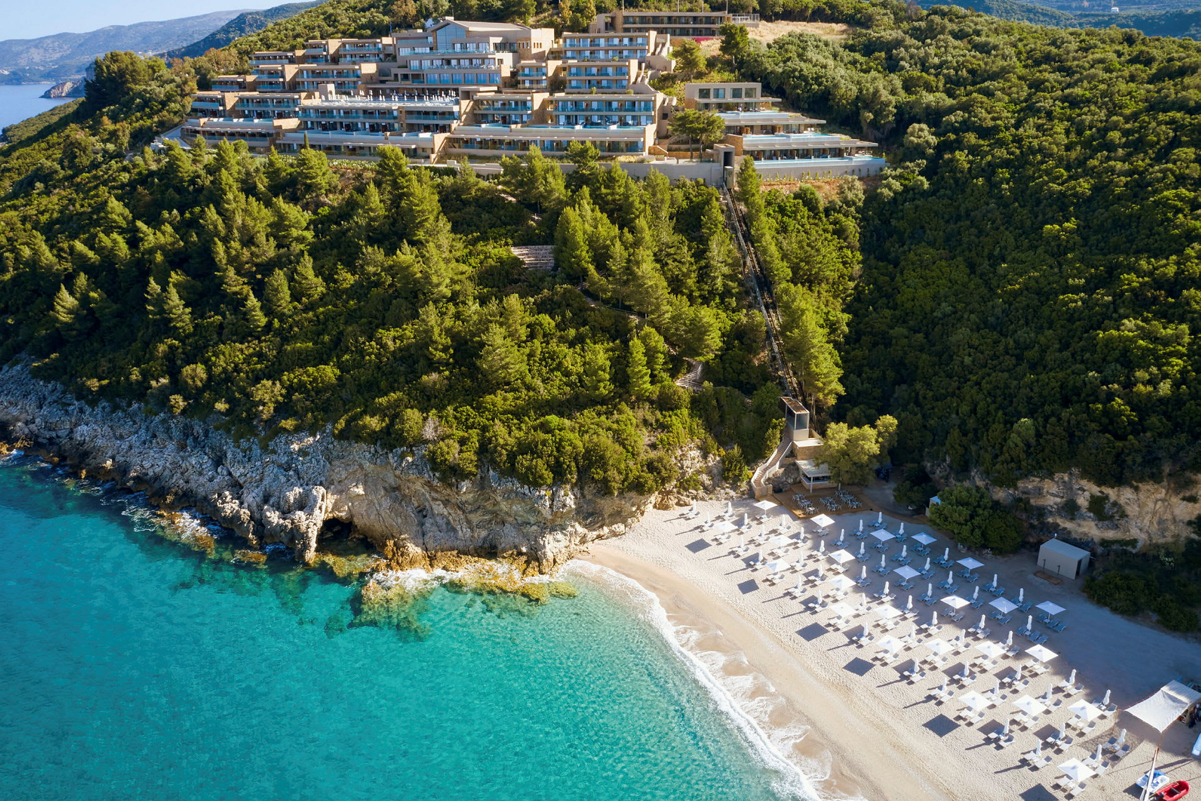 Hotellkomplex på en skogsklädd bergssluttning med utsikt över en strand med parasoller och klart blått hav. (AI-genererad text)