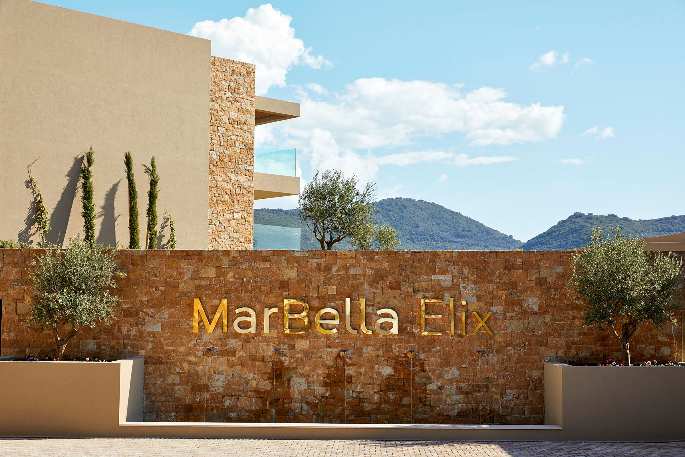 Hotellfasad med texten 'MarBella Elix' på en stenmur, omgiven av träd och berg i bakgrunden. (AI-genererad text)