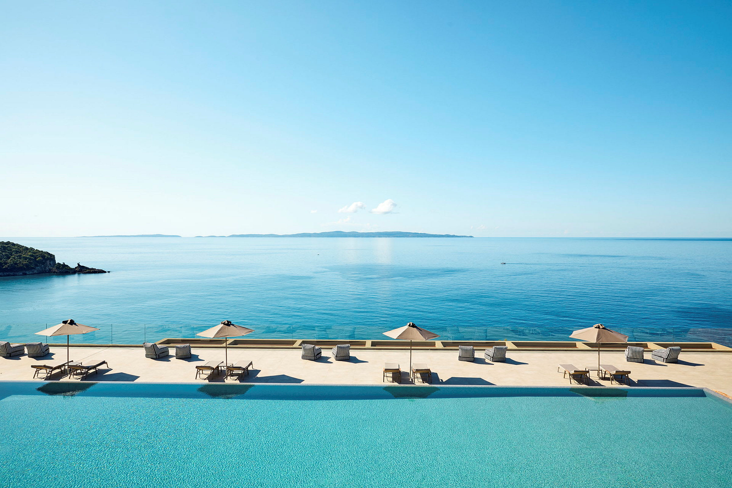 Bild föreställande en infinity-pool med solstolar och parasoll vid en havsutsikt. (AI-genererad text)