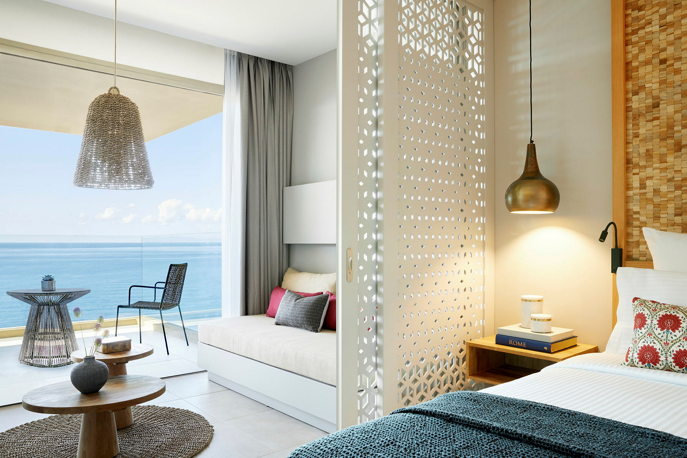 Ett ljust hotellrum med en skjutbar dekorerad avskiljningsvägg, säng och sittgrupp med utsikt över havet. (AI-genererad text)