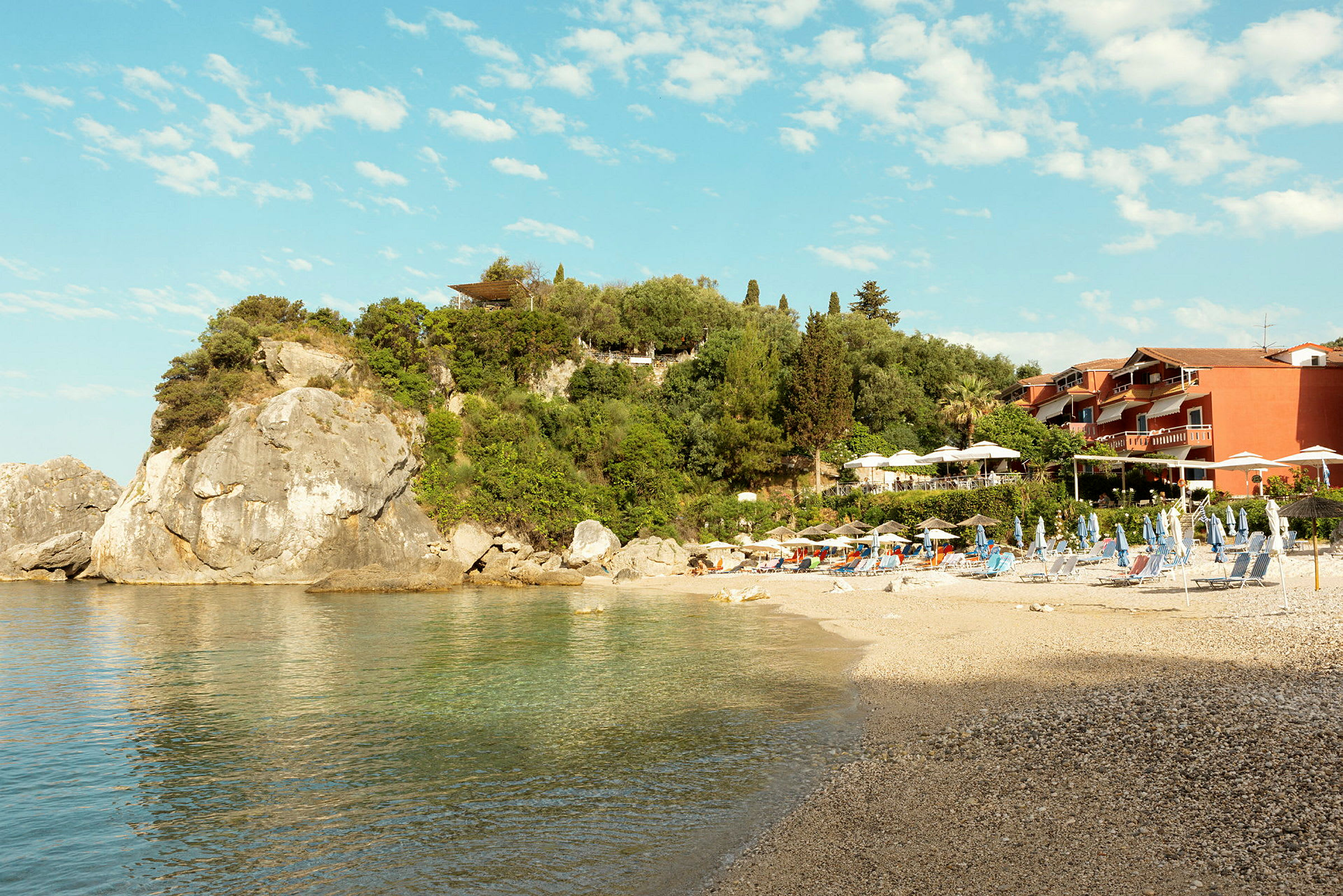En stenig strand intill klart vatten med solstolar och parasoller framför lummig grönska och byggnader. (AI-genererad text)