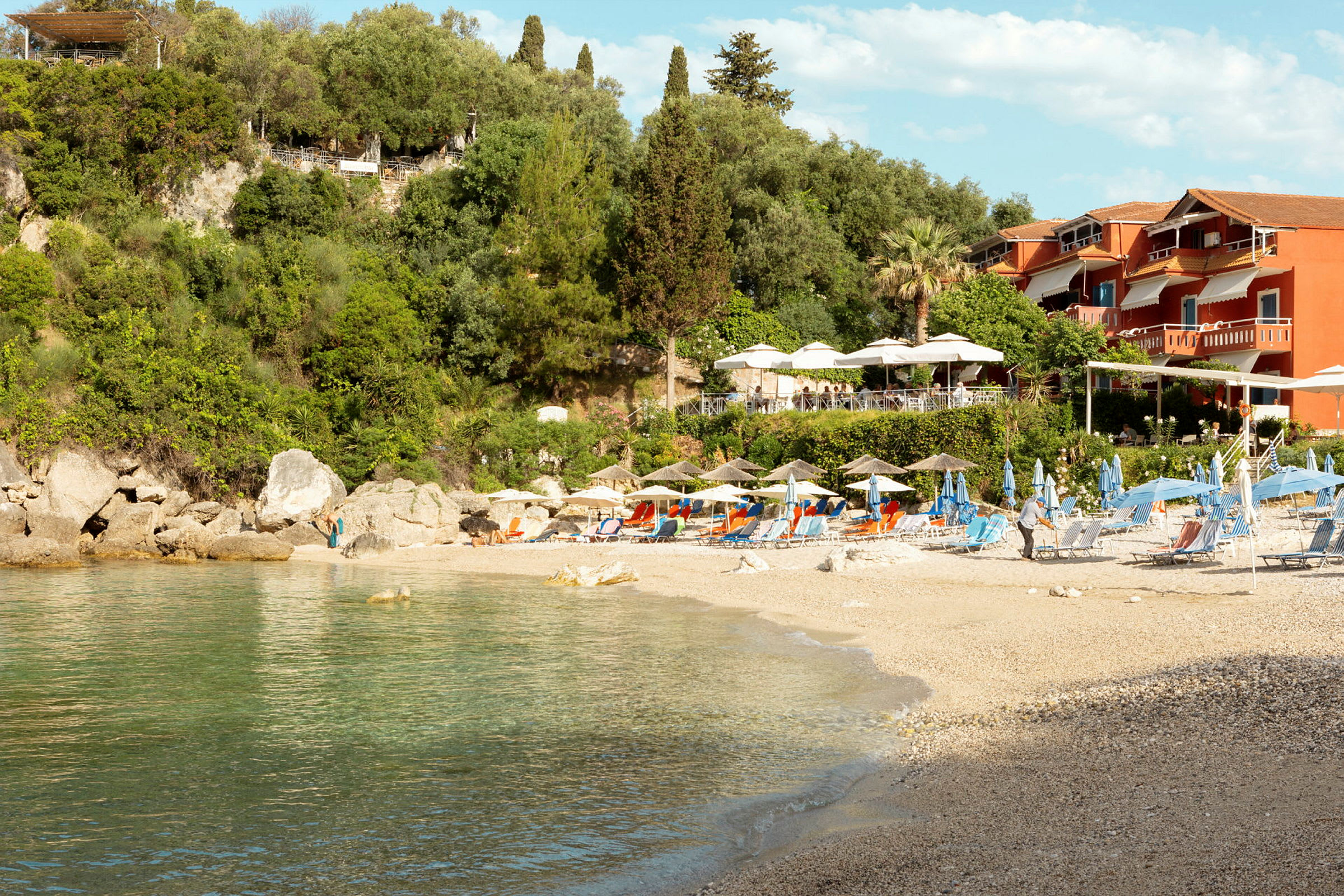 Strand med solstolar och parasoller, grönskande sluttning och röda byggnader i bakgrunden. (AI-genererad text)