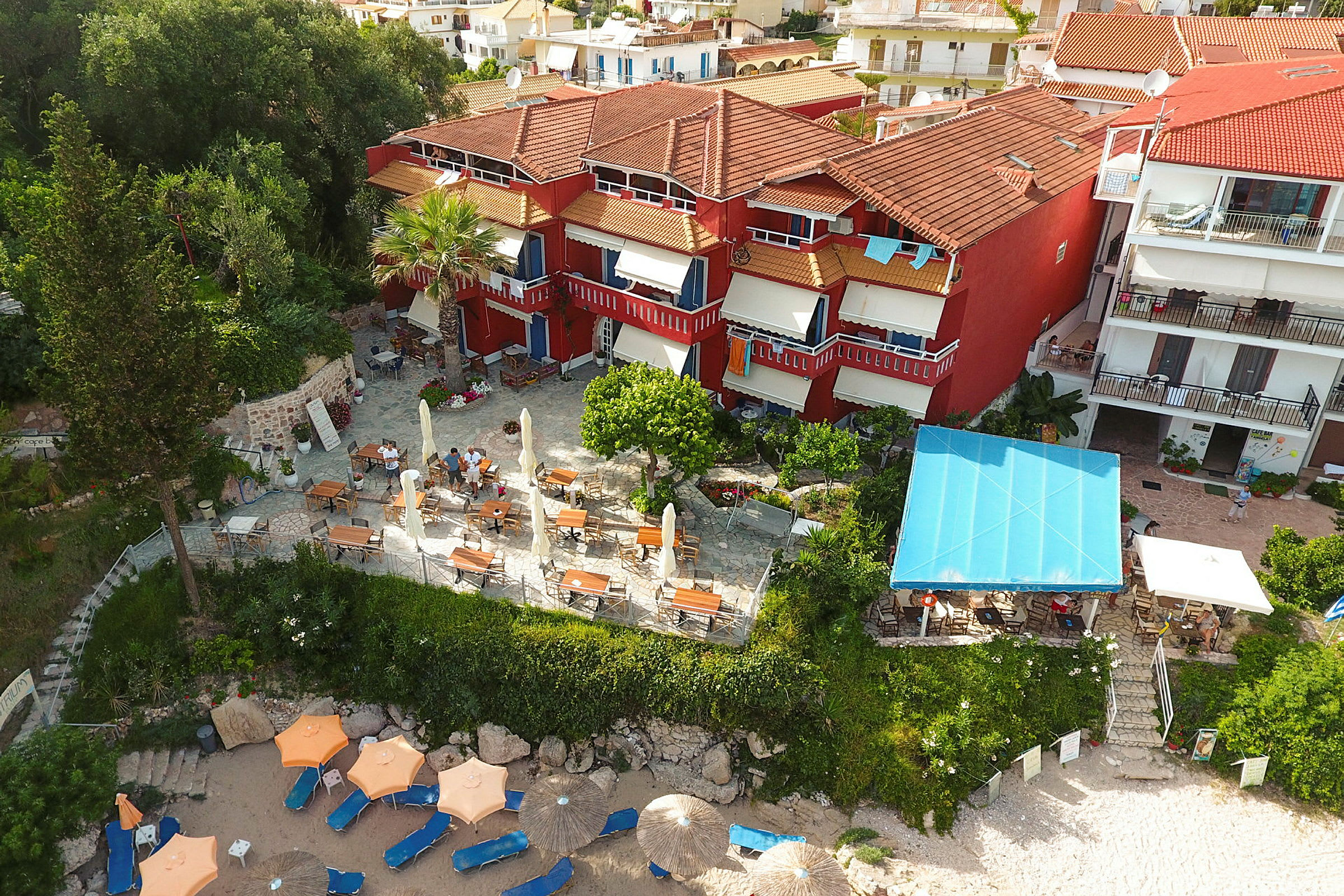 Flygbild över hotell med röda fasader, en terrass med bord och stolar samt parasoller på en sandstrand. (AI-genererad text)